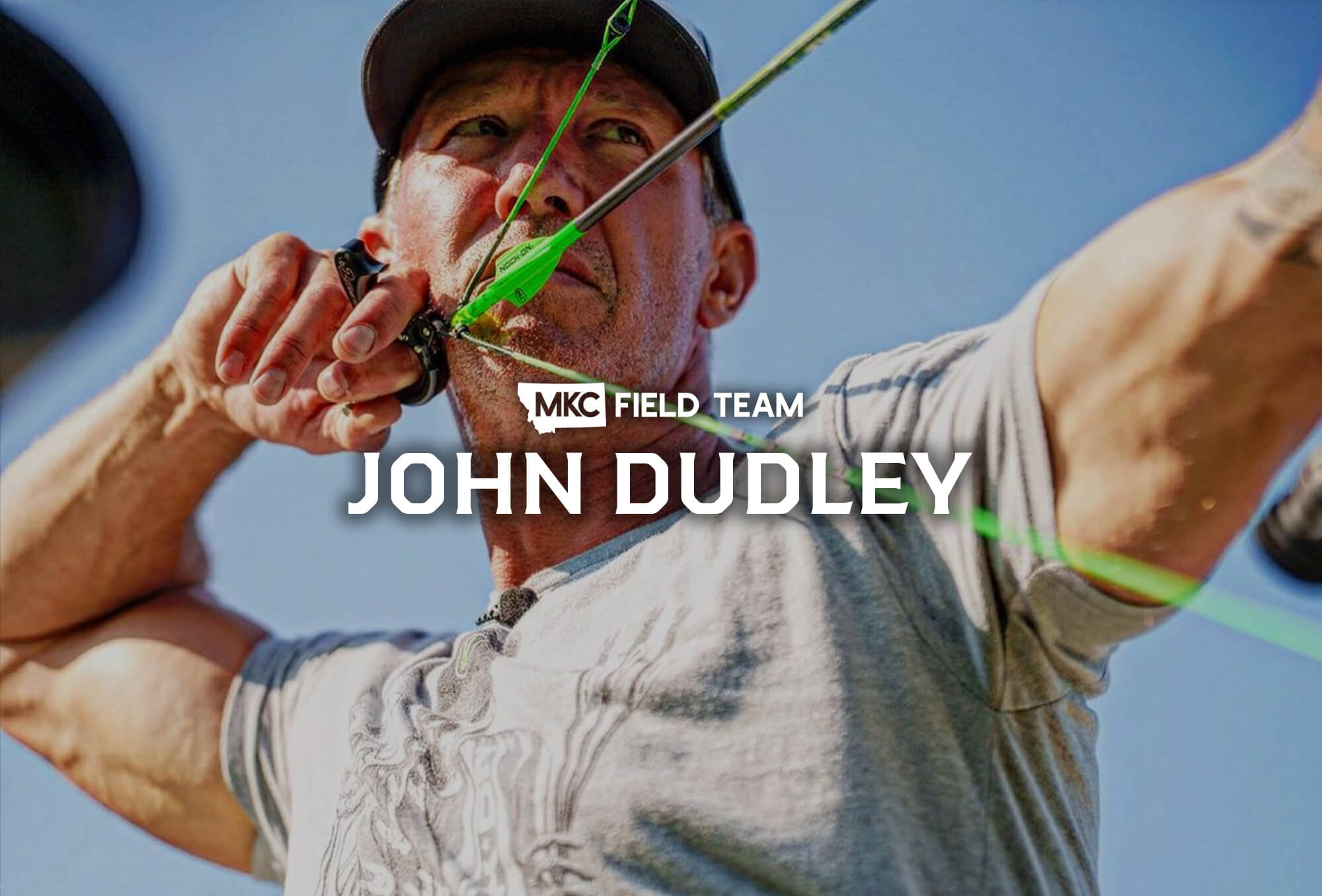 John Dudley stands looking intensely into the distance with an arrow nocked and bow drawn, ready to shoot.