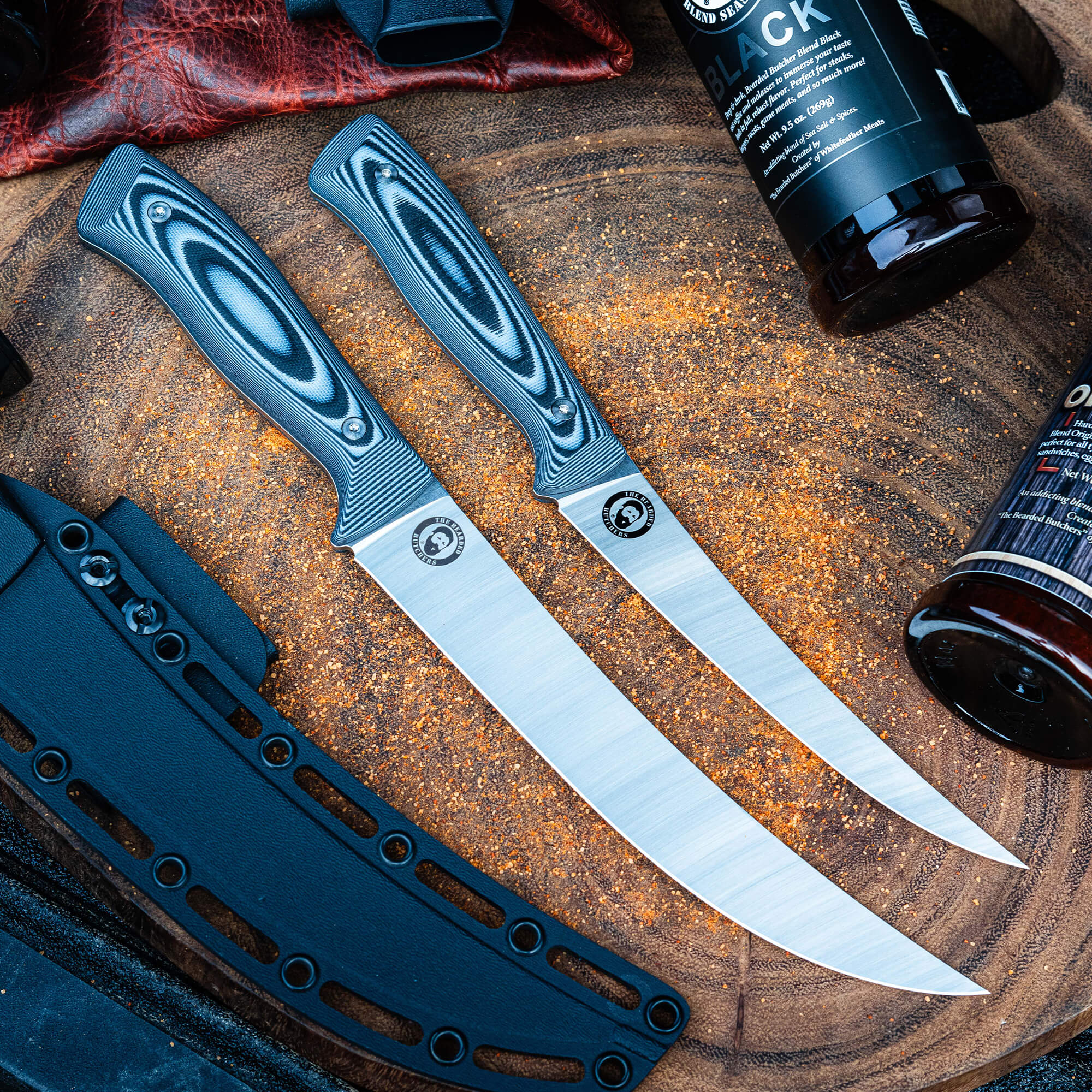 Two high-quality Bearded Butchers breaking butcher knives with black and silver handles, displayed beside seasoning bottles on a rustic wood surface covered with orange spices.
