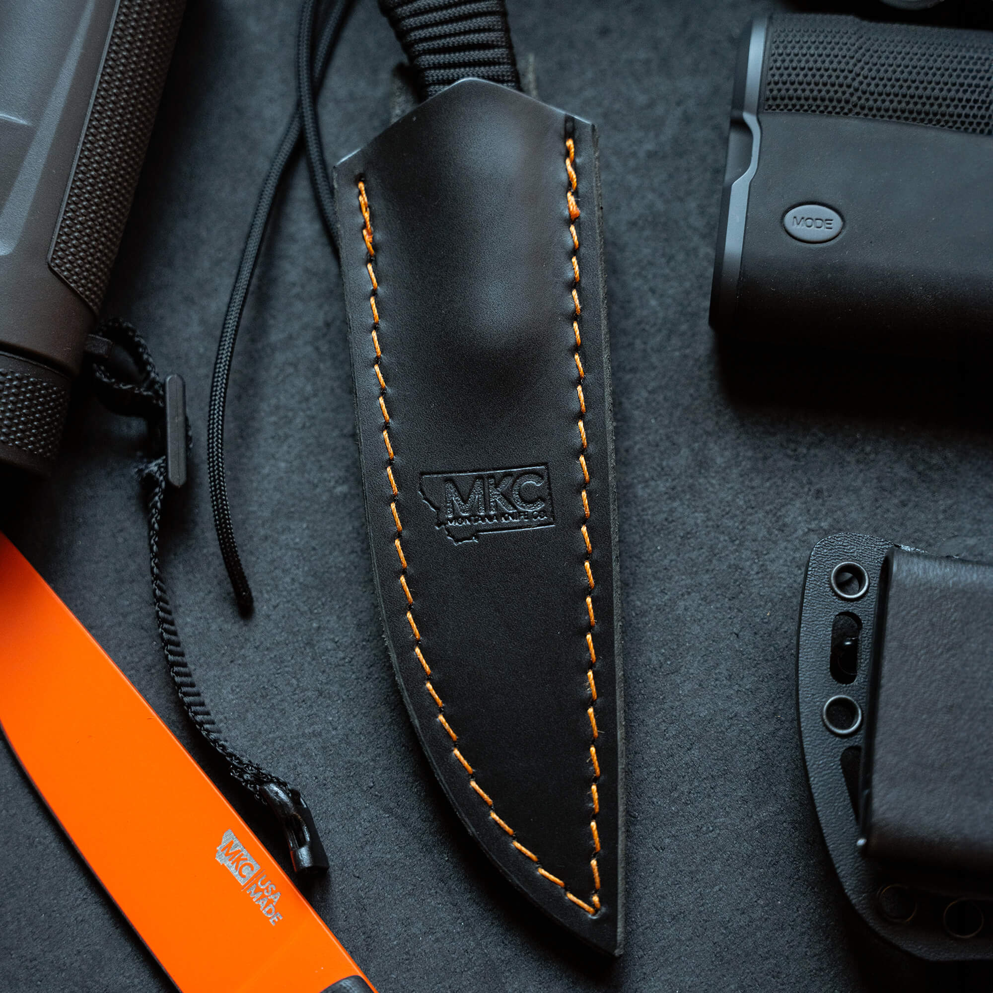 Black leather sheath with orange stitching for vertical belt carry, prominently displaying the MKC logo, featured alongside a black and orange knife handle and other outdoor gear.