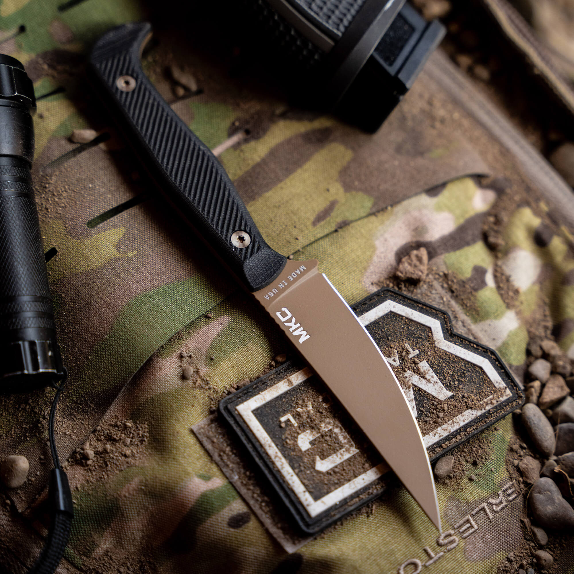 Alt text: Close-up of a KA-BAR 'Battle Goat' tactical knife in Coyote/Black color, displayed on a camouflage background alongside tactical gear, highlighting the sharp, pointed blade and textured handle.