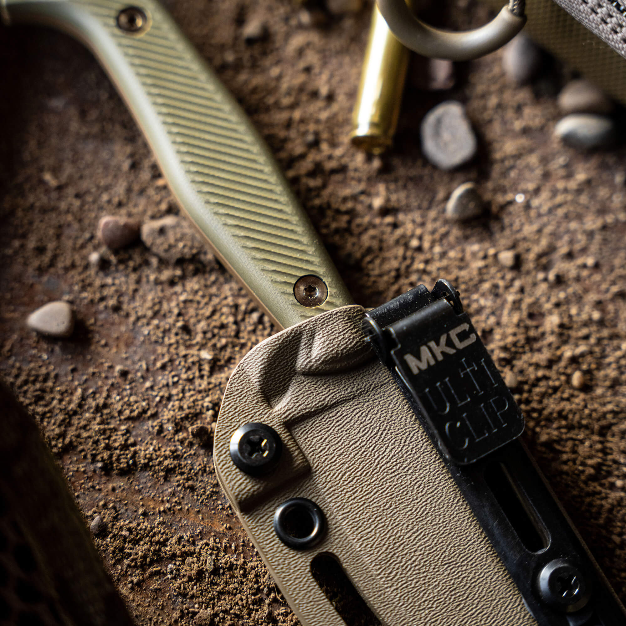 Close-up view of a Battle Goat tactical knife in coyote olive drab, featuring a textured handle and sheath, partially buried in gritty soil with bullets nearby.