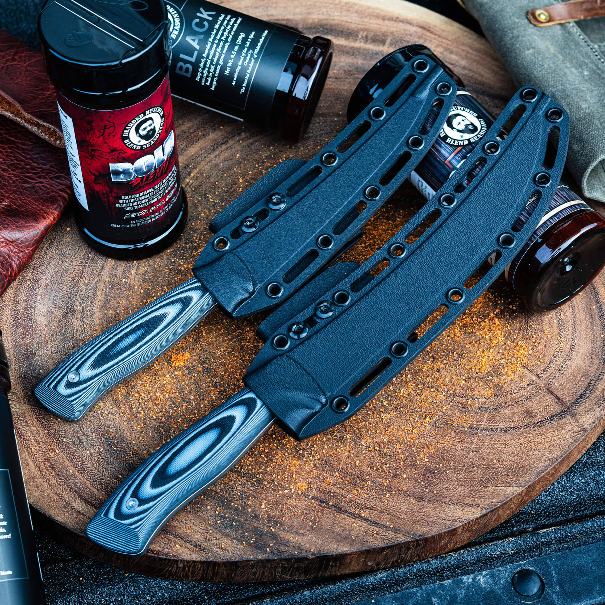 Two Bearded Butchers breaking butcher knives displayed with sheaths on a wooden board alongside Black Bearded Butchers Blend seasoning bottles, set against a rustic background with a hint of sparkle.