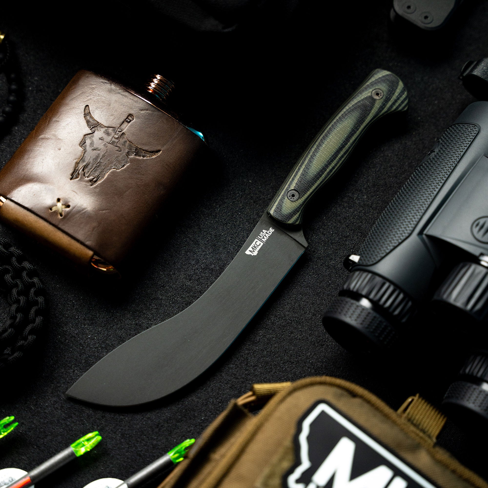 Alt Text: "Beartooth Pro Skinning Blade with a green and black handle displayed against a backdrop of tactical gear including a flint-brown leather flask with bull logo, black binoculars, and assorted hunting equipment."