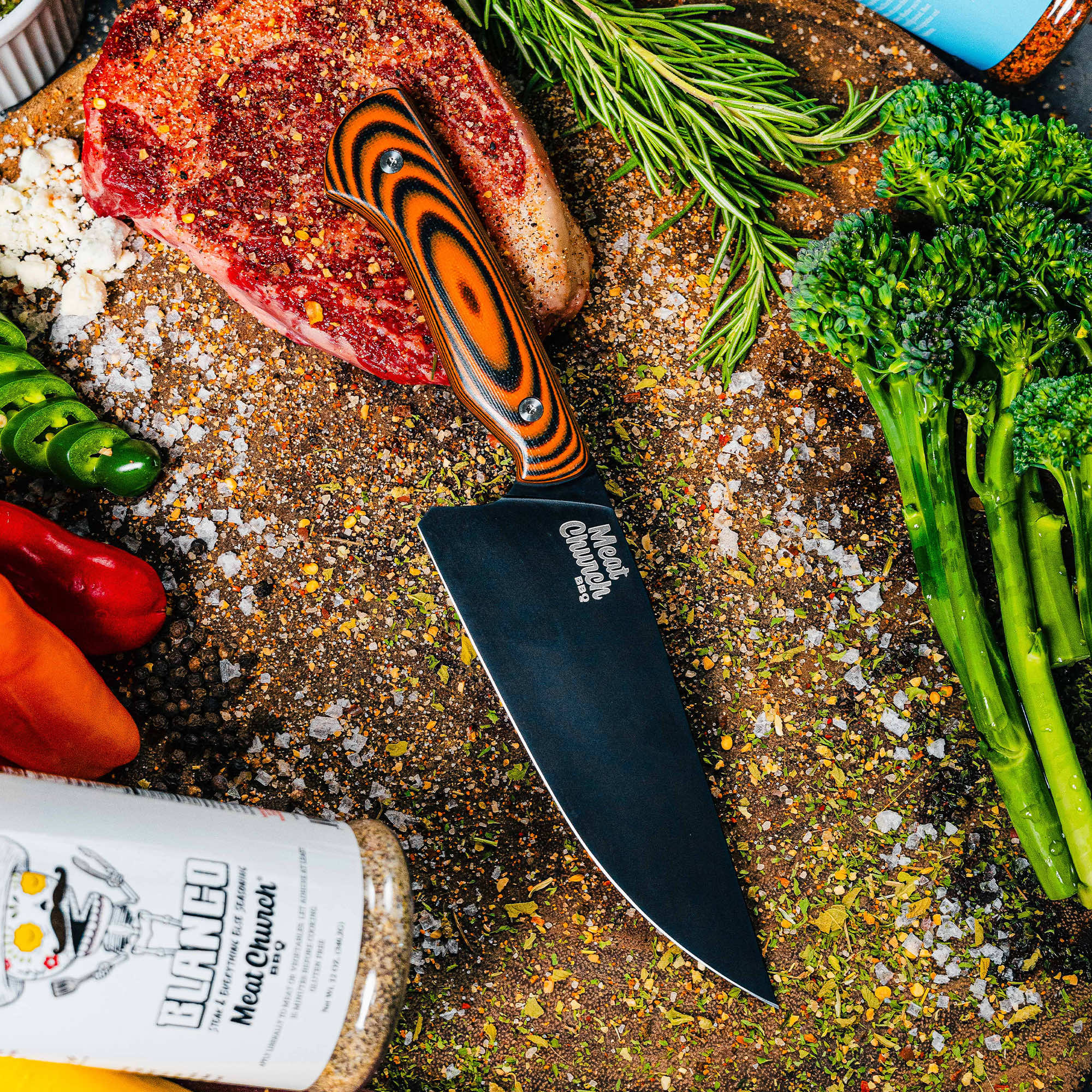 Meat Church chef knife on a wooden cutting board surrounded by fresh vegetables and seasonings, showcasing its sleek design.