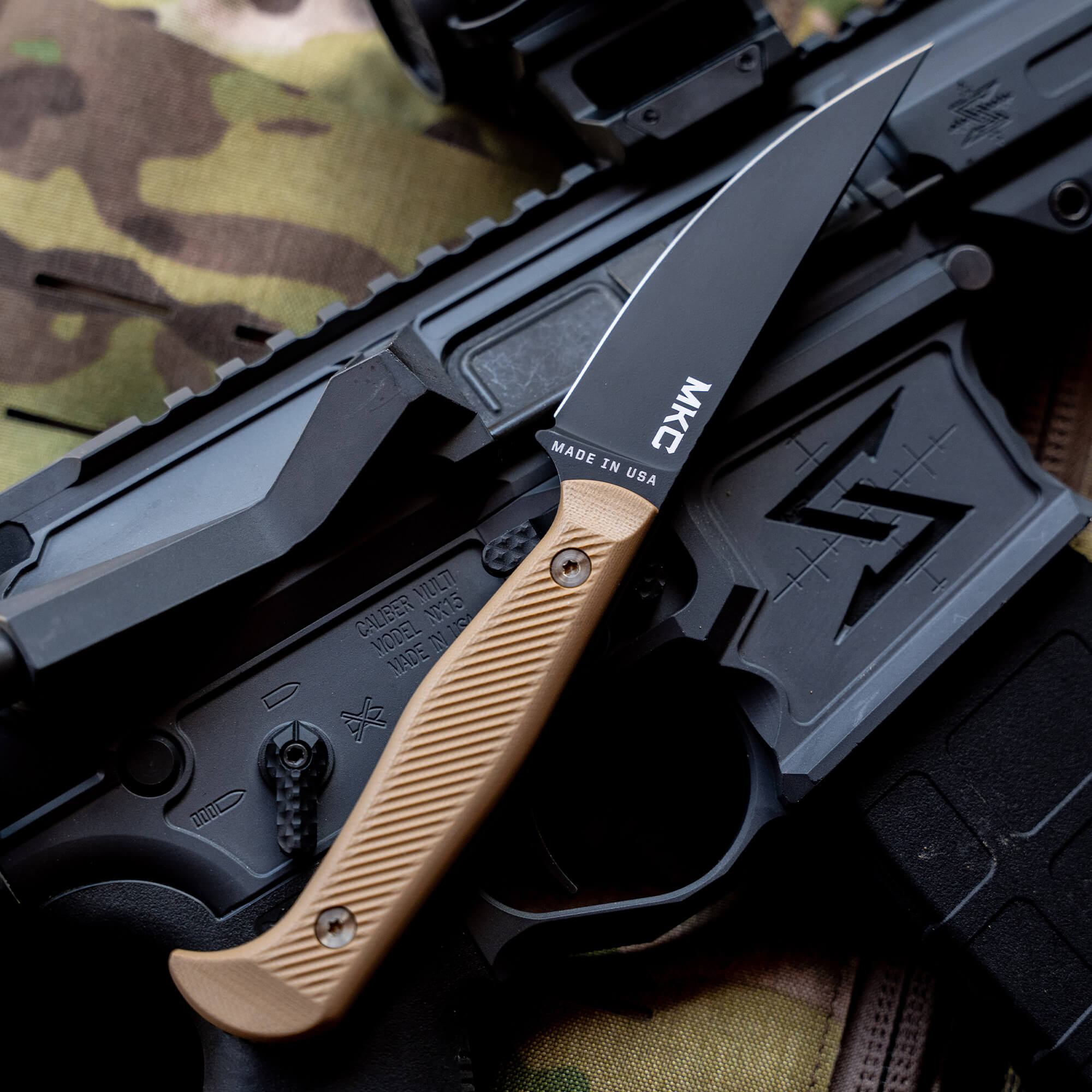 Alt text: "Close-up view of a Battle Goat tactical knife in black and coyote tan colors, resting on a camouflage background and an AR-15 style rifle marked 'Made in USA'."