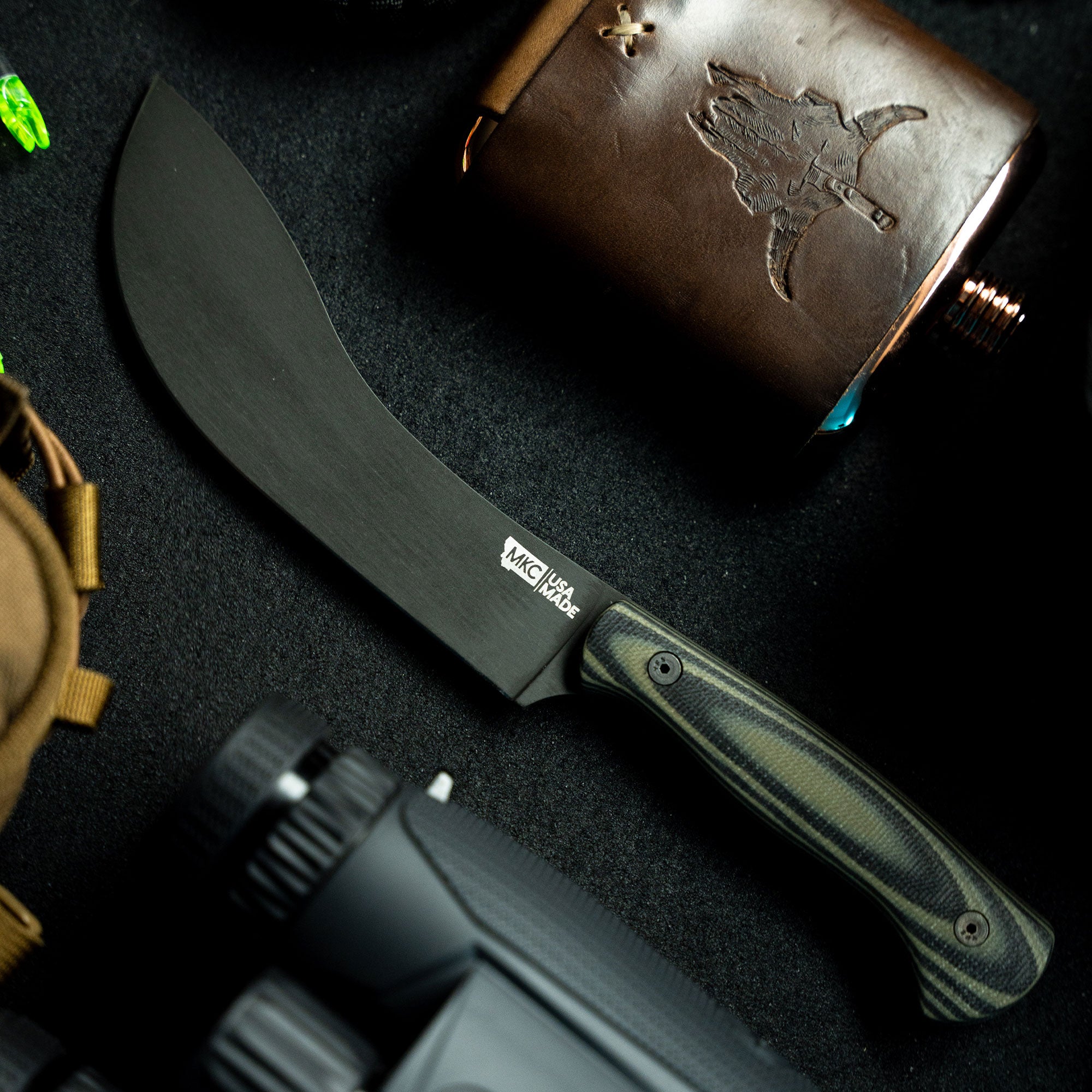 Alt text: "Close-up of a Beartooth Pro Skinning Blade, featuring a curved matte black blade and a green and black striped handle. The knife is displayed against a dark background with a leather flask embossed with a map and other outdoor