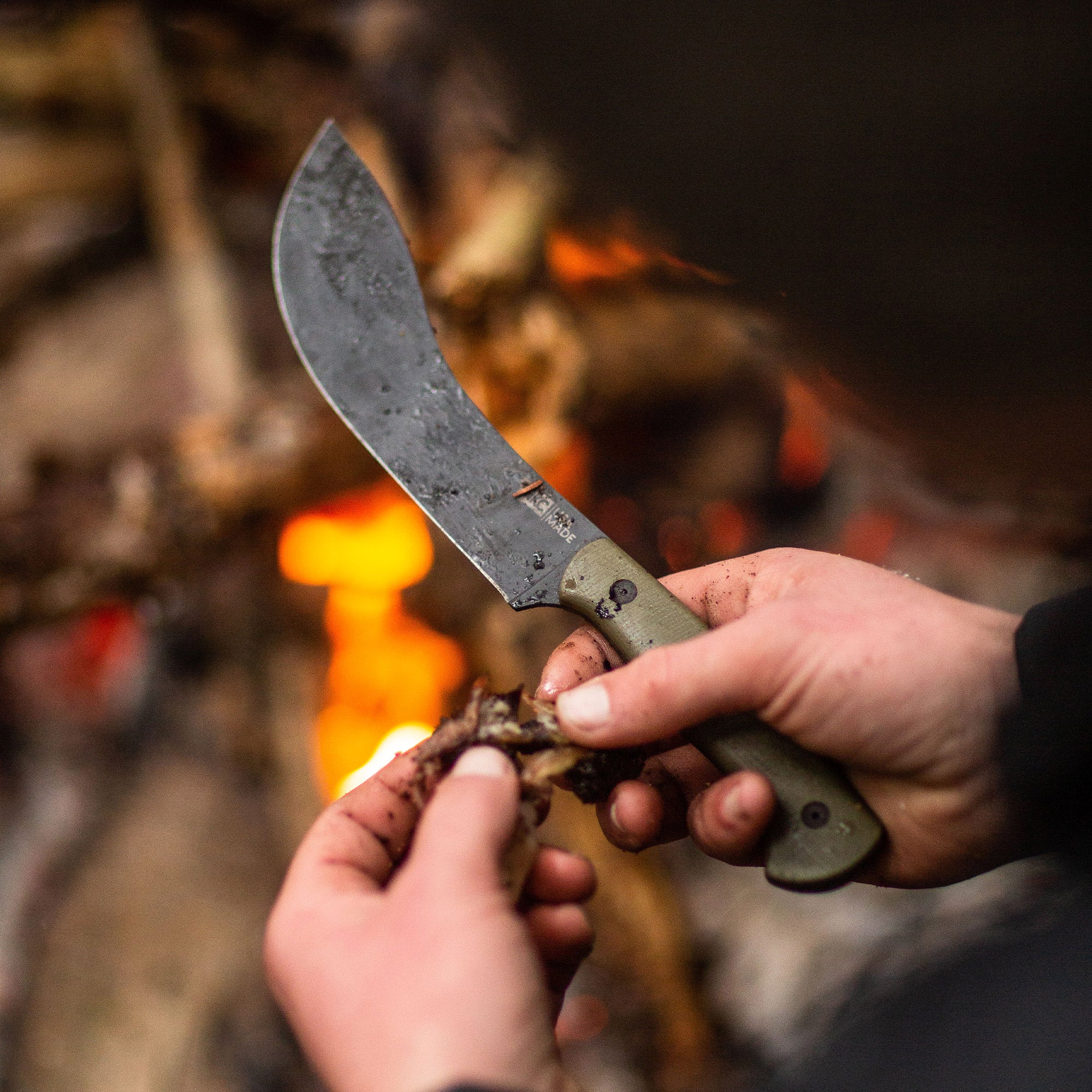 Close-up of a person using the Beartooth Pro Skinning Blade with an olive handle to skin game near a campfire.