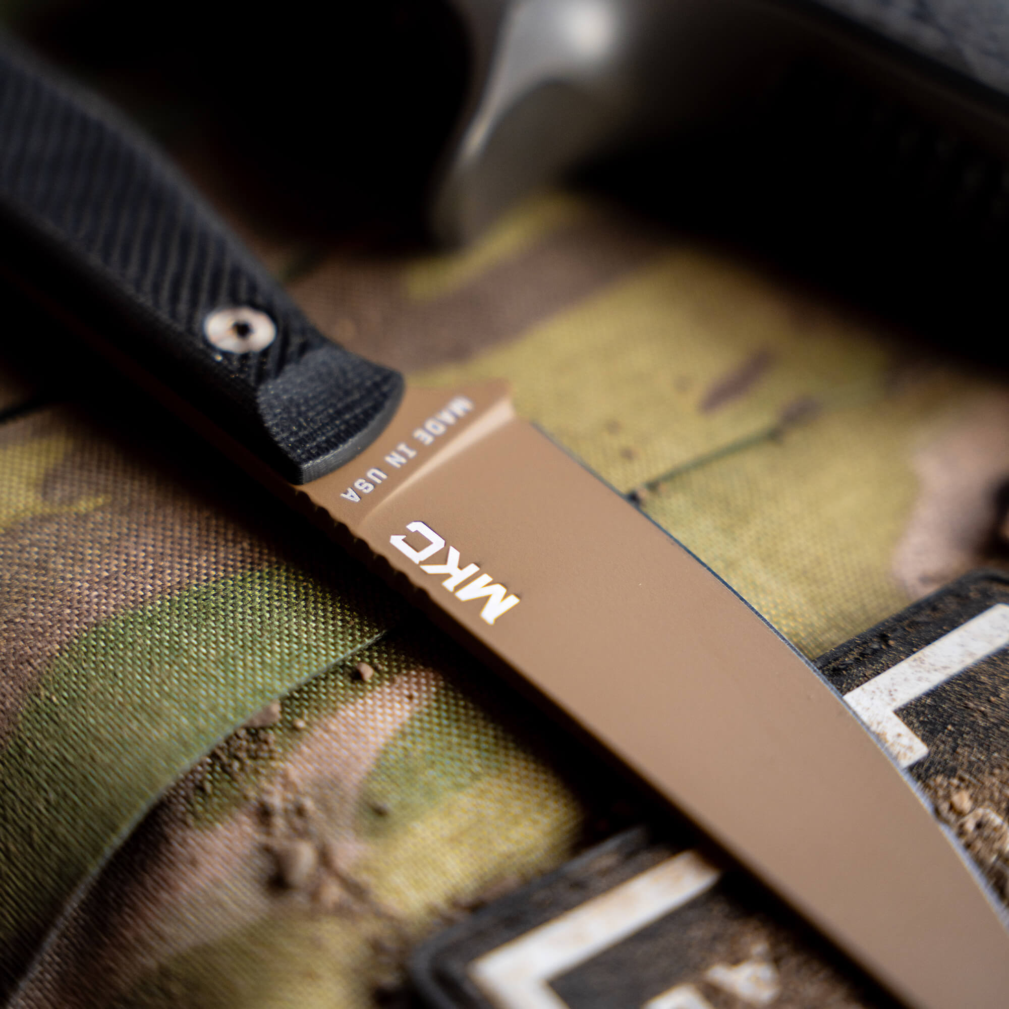 Close-up of a Battle Goat tactical knife in coyote black colorway, featuring a brown blade with the “MADE IN USA” inscription, resting on a camouflaged surface.
