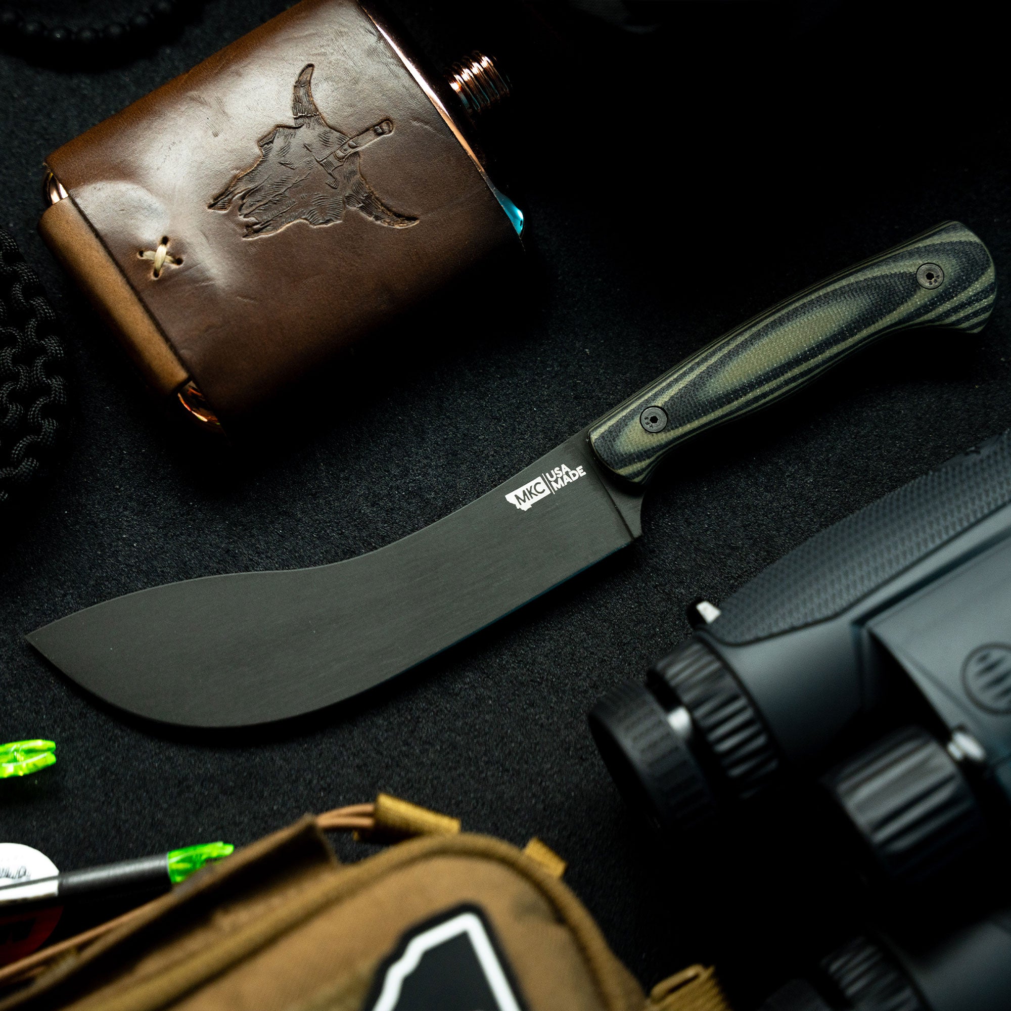 "Beartooth professional skinning knife with a curved black blade and green and black striped handle, displayed on a dark background next to a leather flask with a carved stag design."