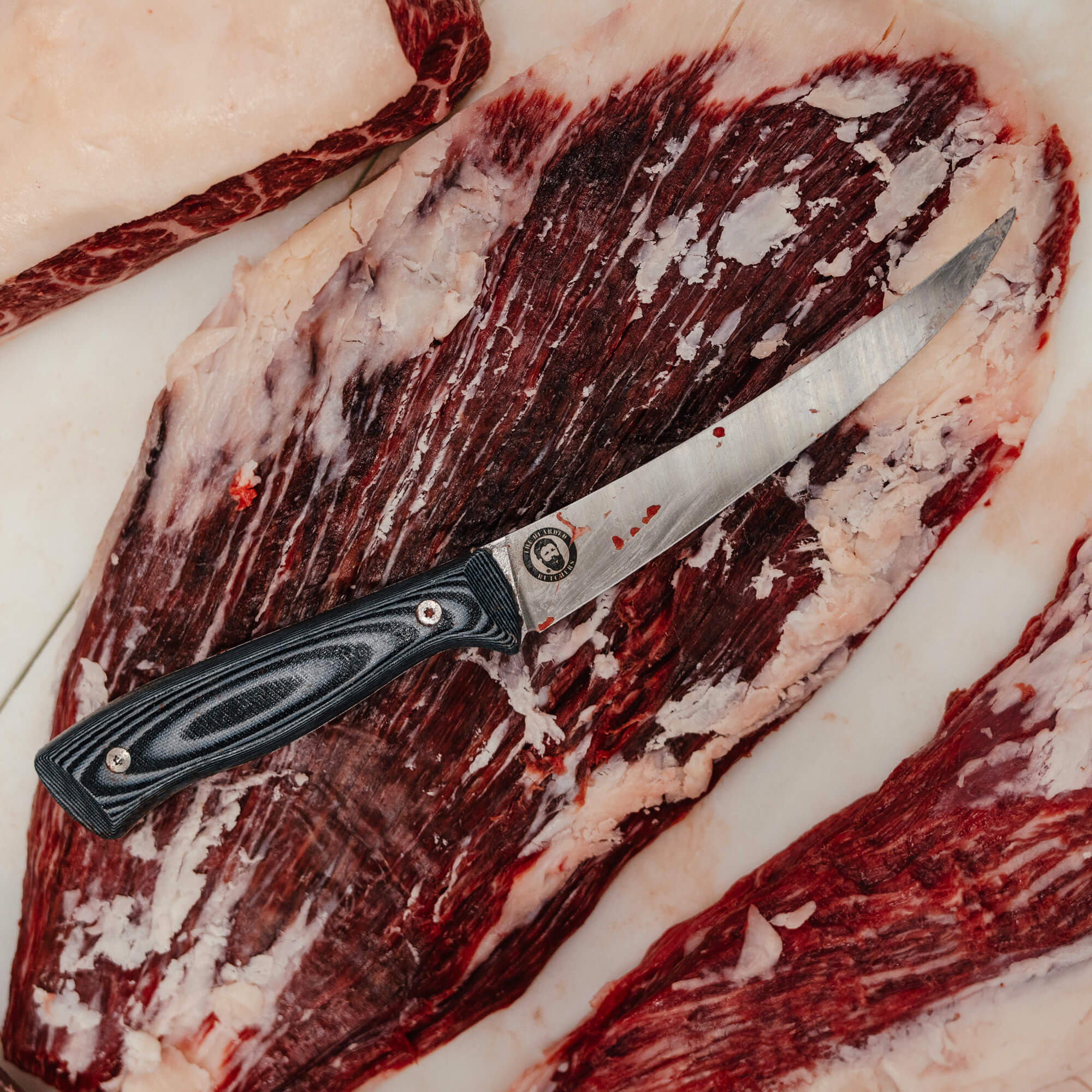 Alt text: "Bearded Butchers boning butcher knife with a dark handle, featured on a background of raw marbled meat, emphasizing the knife's sharp blade and professional quality."