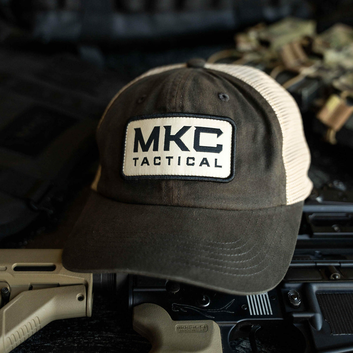 MKC Tactical Classic Trucker hat featuring a black and white logo on a brown and tan cap, placed on tactical gear.