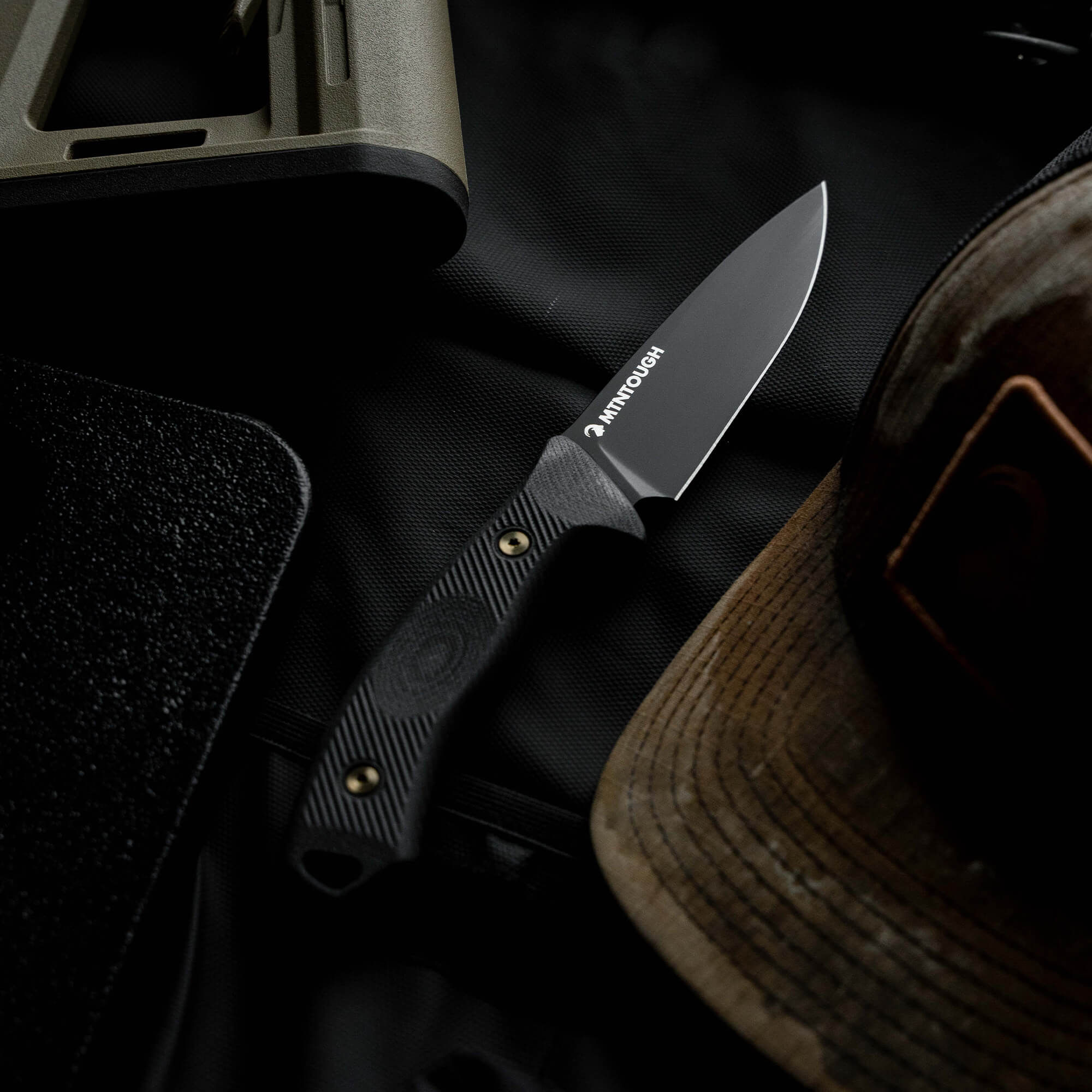 MTNTOUGH TF24 knife with a black blade and textured handle, resting on a dark background with gear and a hat.