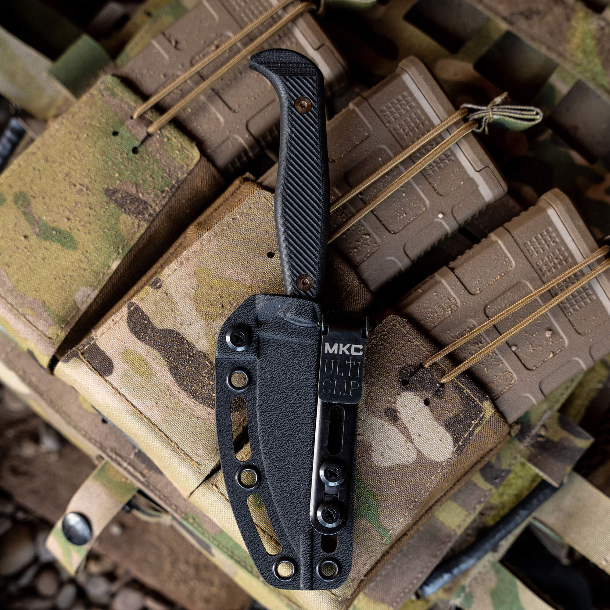 Alt text: "Black BATTLE GOAT tactical knife secured in a black Kydex sheath with an MKC UltiClip, mounted on a camouflage tactical vest featuring various pouches and straps."