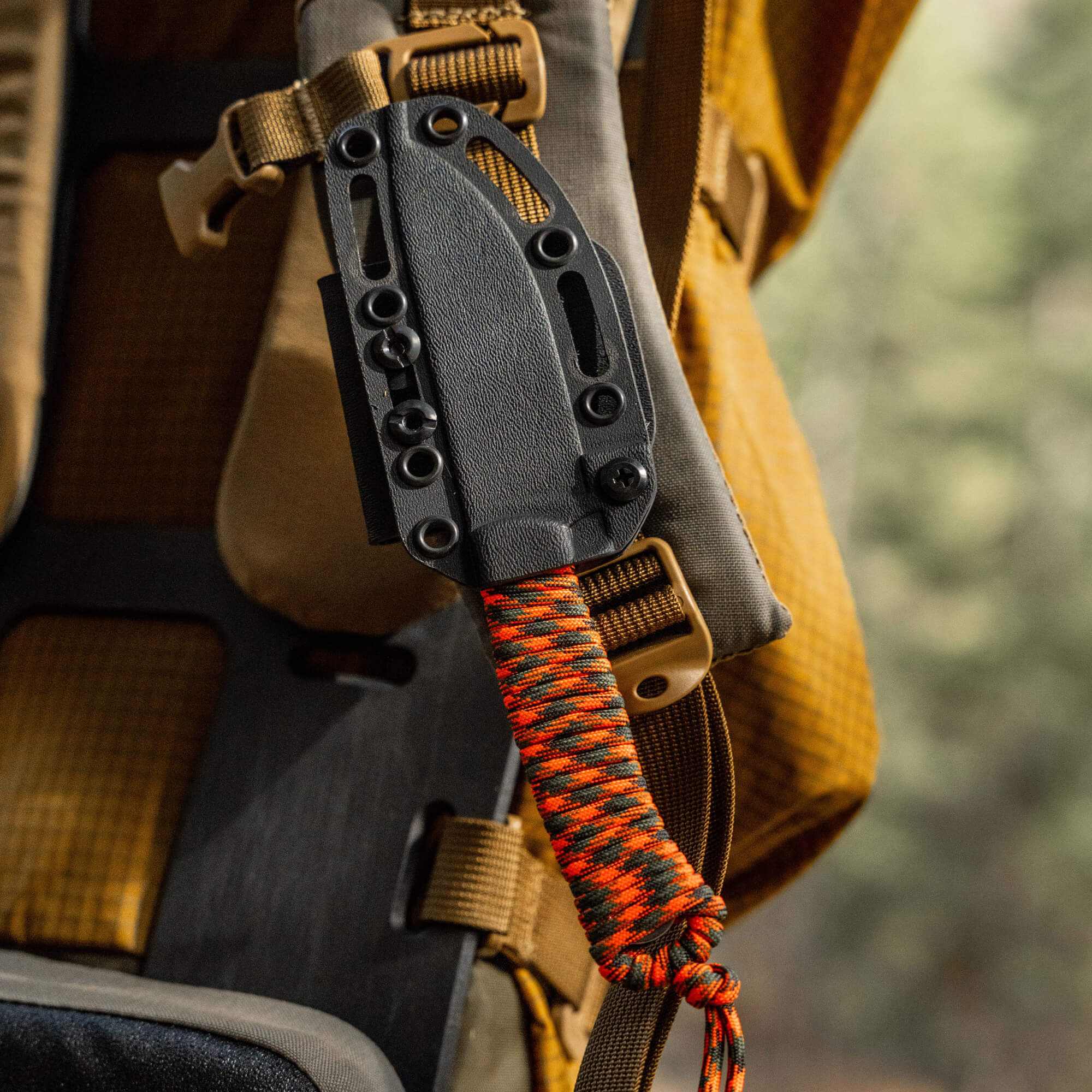 Alt text: Close-up of an ElkShape MagnaCut Stoned Goat 2.0 knife sheathed and attached to a tactical backpack with an orange and black paracord, emphasizing its rugged outdoor design.