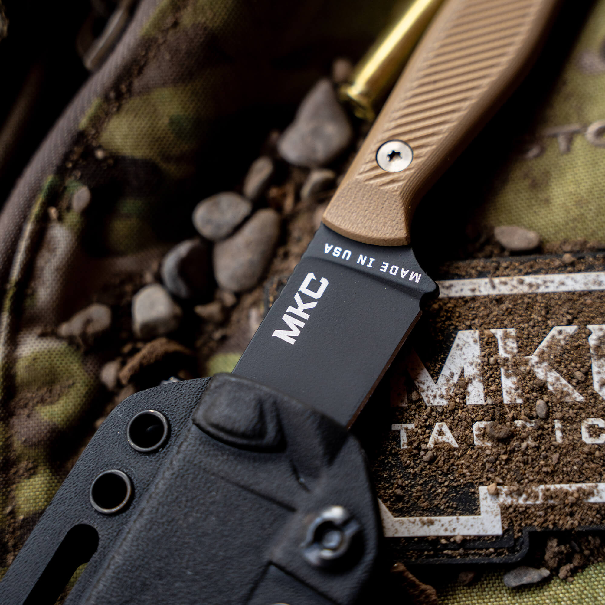 Close-up of a tactical knife, the MKC Battle Goat in black and coyote color, partially sheathed in a black holster, resting on a camouflage background with pebbles.