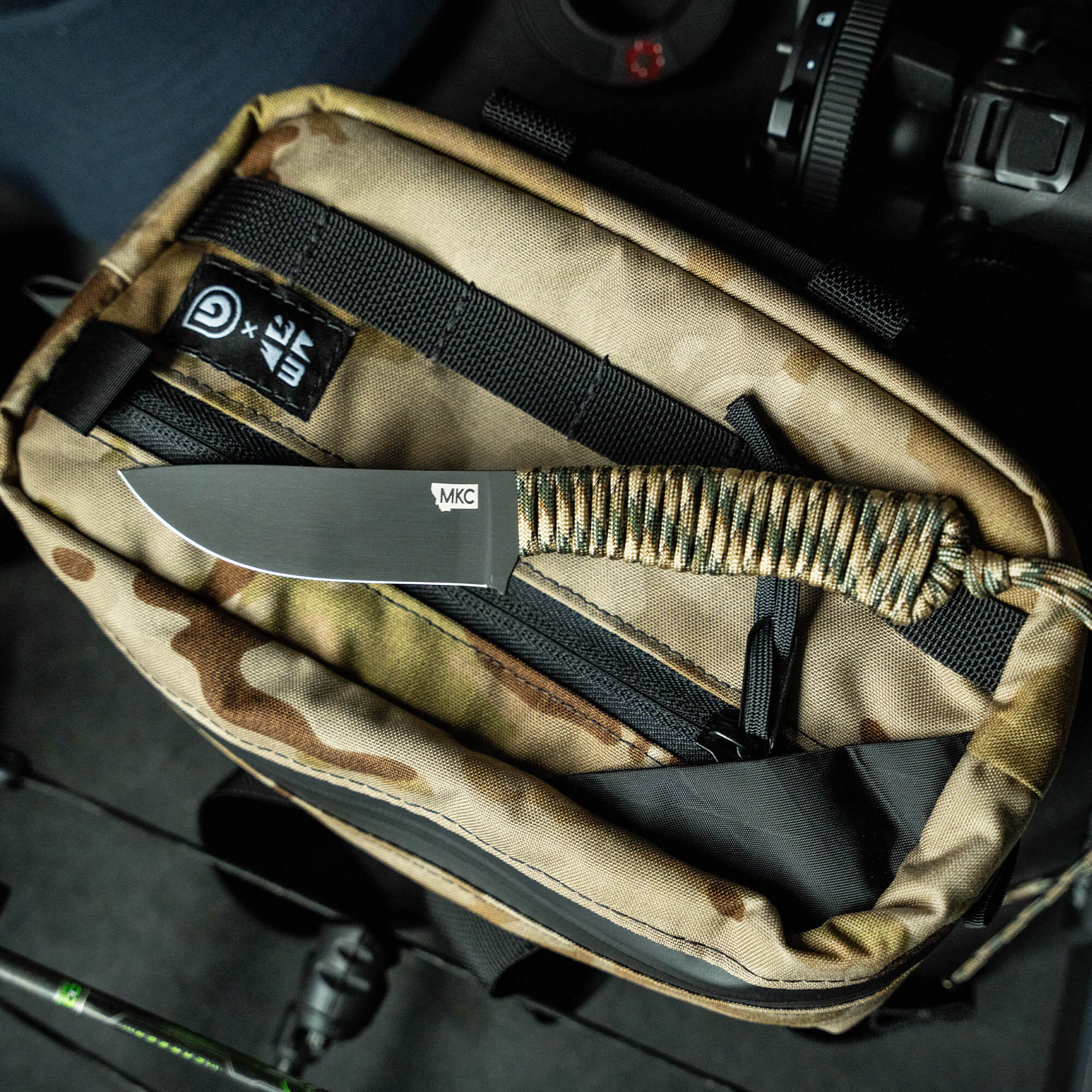 Alt text: Tactical hunting knife with olive green paracord-wrapped handle and polished steel blade, displayed inside an open camouflaged tactical pouch, with dark modern equipment in background.