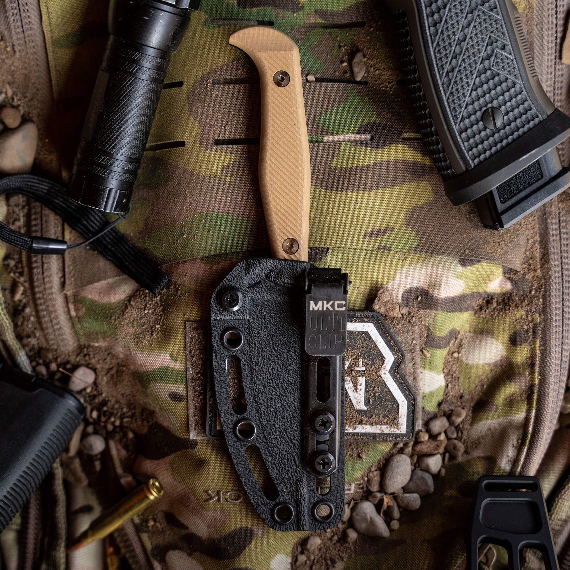 Alt text: "Close-up view of the BATTLE GOAT tactical knife in black and coyote tan, featuring a tan handle and black sheath, secured on a camouflaged military backpack alongside various tactical gear."
