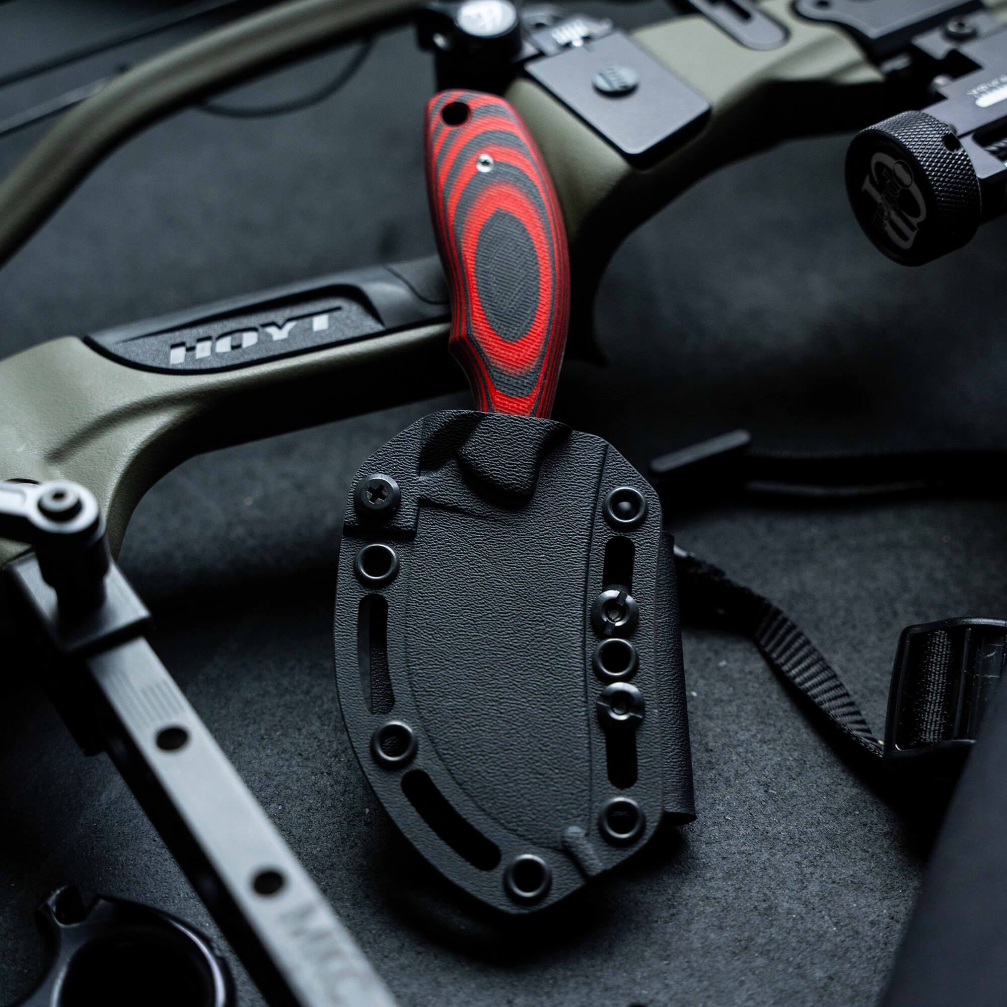 Red and black handled Packout Skinner knife with a black sheath, displayed against a backdrop of archery equipment, including a Hoyt bow.