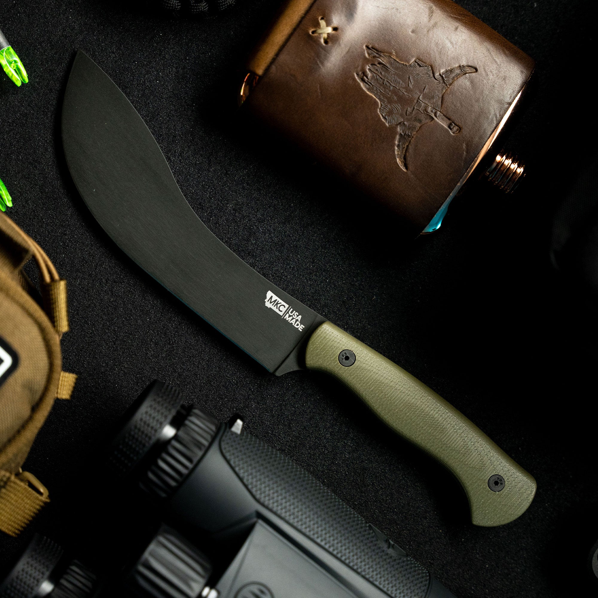 Alt text: "Beartooth Pro Skinning Blade with olive green handle, prominently displayed alongside hunting gear, including a flint flask with etched map design, camera, and binoculars on a dark textured surface."