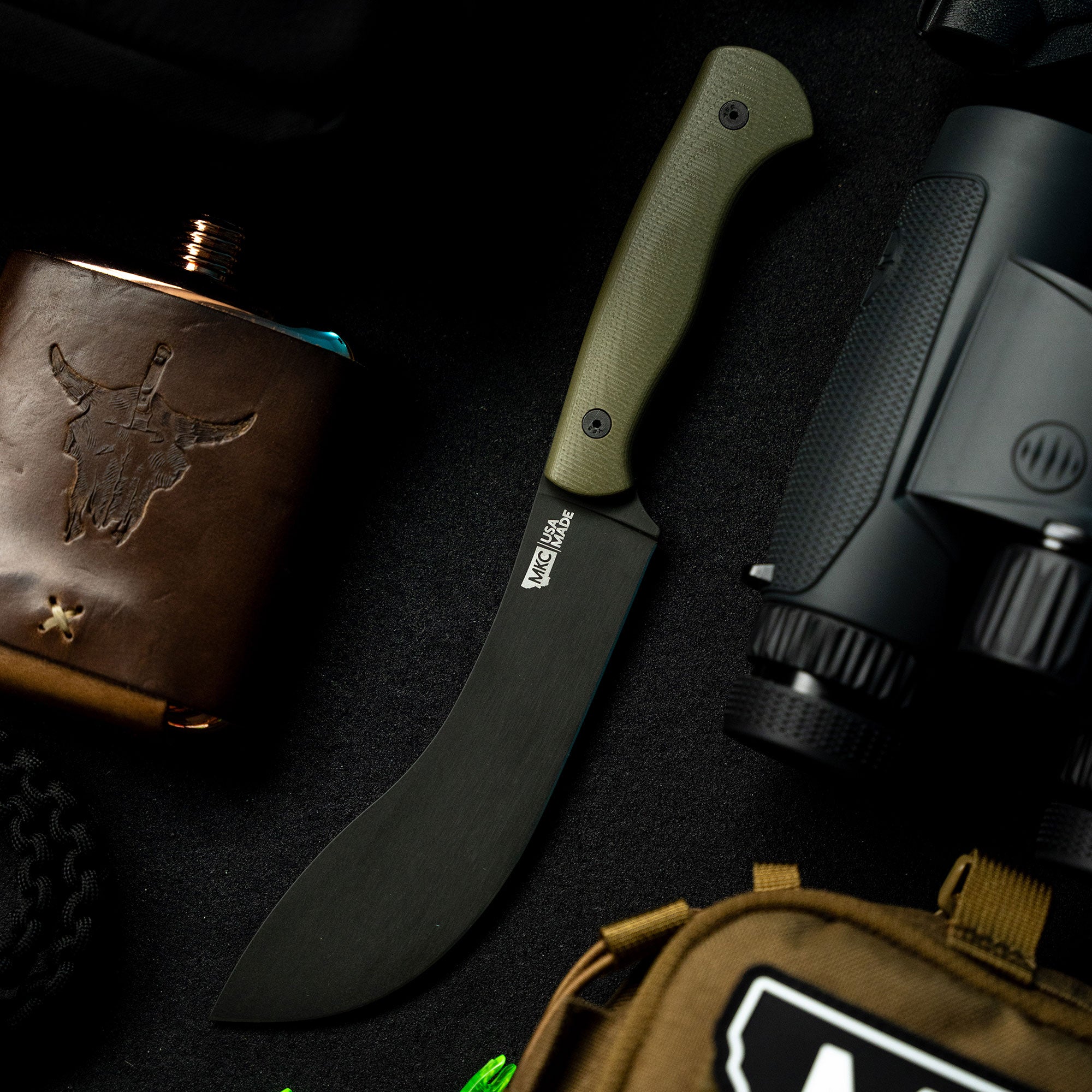 Alt text: "BEARTOOTH Pro Skinning Blade with olive green handle and matte black blade, displayed alongside a brown leather flask and black binoculars on a black textured background."
