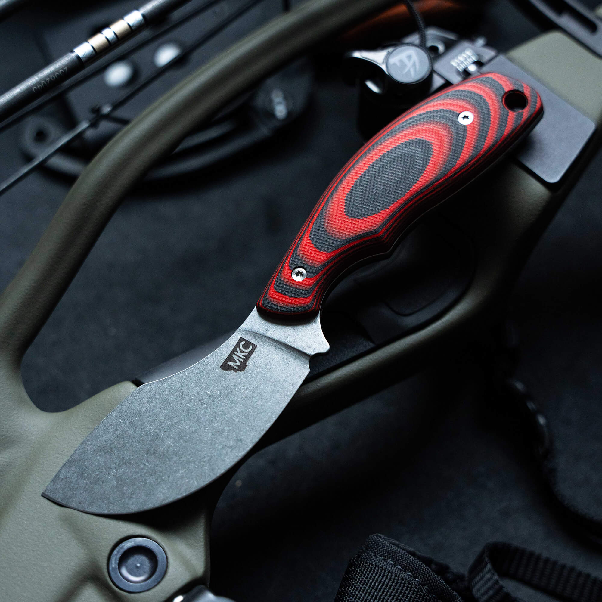 Alt text: "Close-up view of a PACKOUT Skinner knife with a red and black ergonomic handle and a rugged matte-finish blade, displayed on a tactical background with parts of firearms visible, indicating the knife's sturdy, outdoor and tactical use."