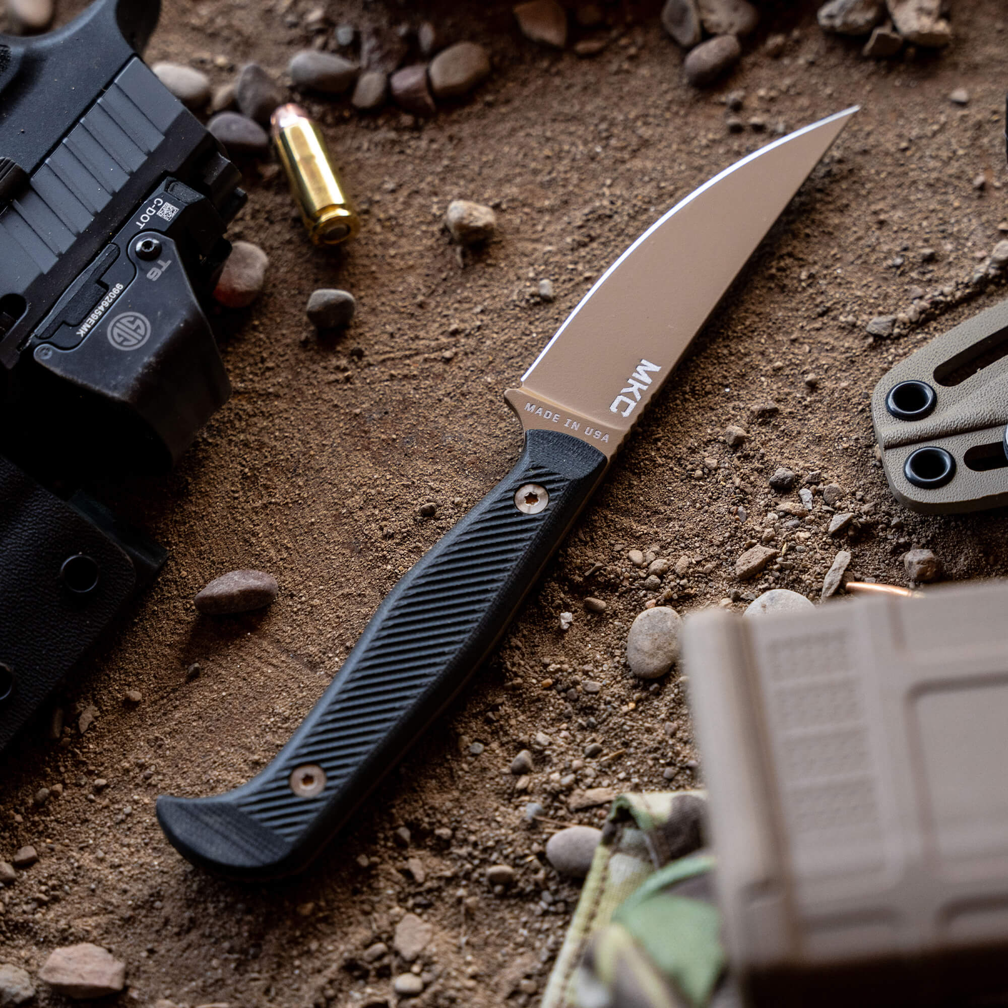 Alt text: "A high-quality Winkler Battle Goat fixed blade knife with a matte silver blade and black textured handle, displayed on a gritty sandy surface surrounded by a handgun, empty bullet casings, and tactical gear, highlighting its utility in rugged