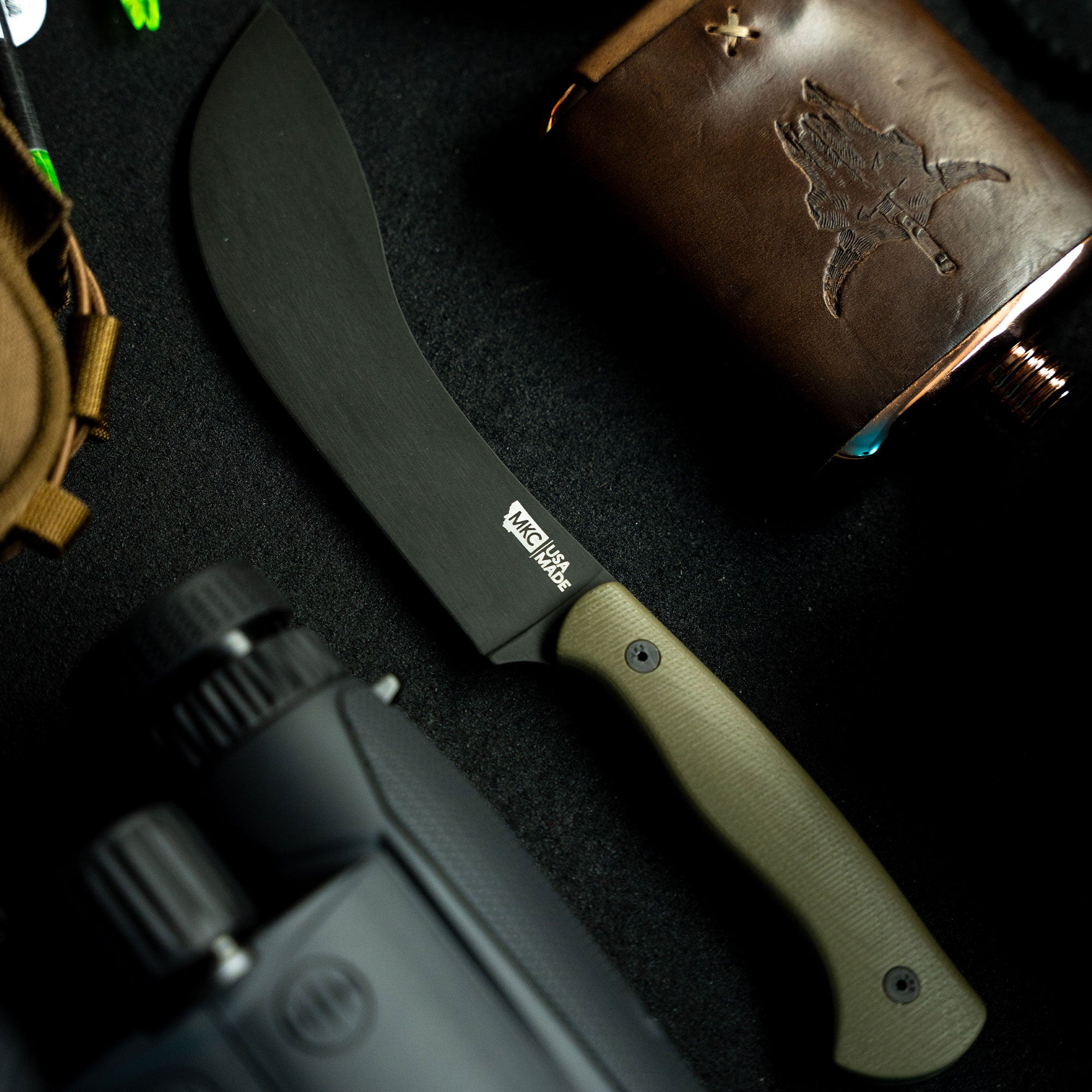 Alt text: "Close-up of Beartooth Pro Skinning Blade with olive handle, featuring a curved black blade, displayed on a dark background with a leather pouch and outdoor gear."