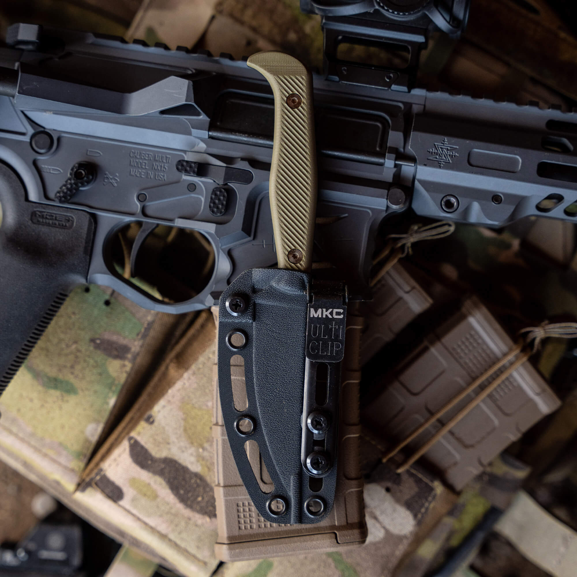 Alt text: Close-up of a tactical knife with a tan handle attached to a black and gray modern sporting rifle, featuring various accessories and markings. The knife is secured in a black sheath with visible adjustment screws, attached to the rifle's M-