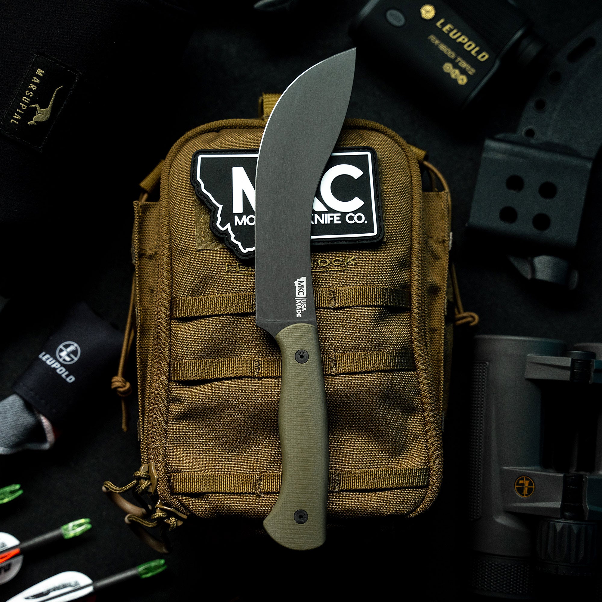 Alt text: "Beartooth Pro Skinning Blade with an olive green handle displayed against a tactical gear bag with outdoor equipment and various logos, highlighting its robust design suitable for hunting and outdoor activities."