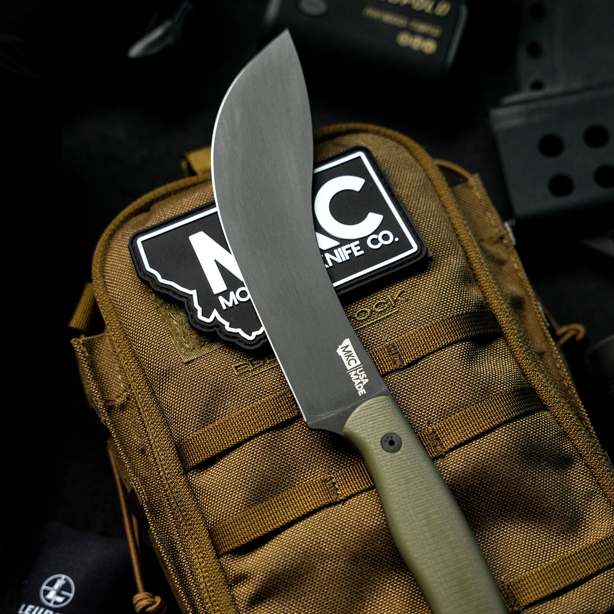 Alt text: Close-up view of a Beartooth Pro Skinning Blade with an olive handle, prominently displayed against a tactical backpack, highlighting its sleek design and sharp edge.