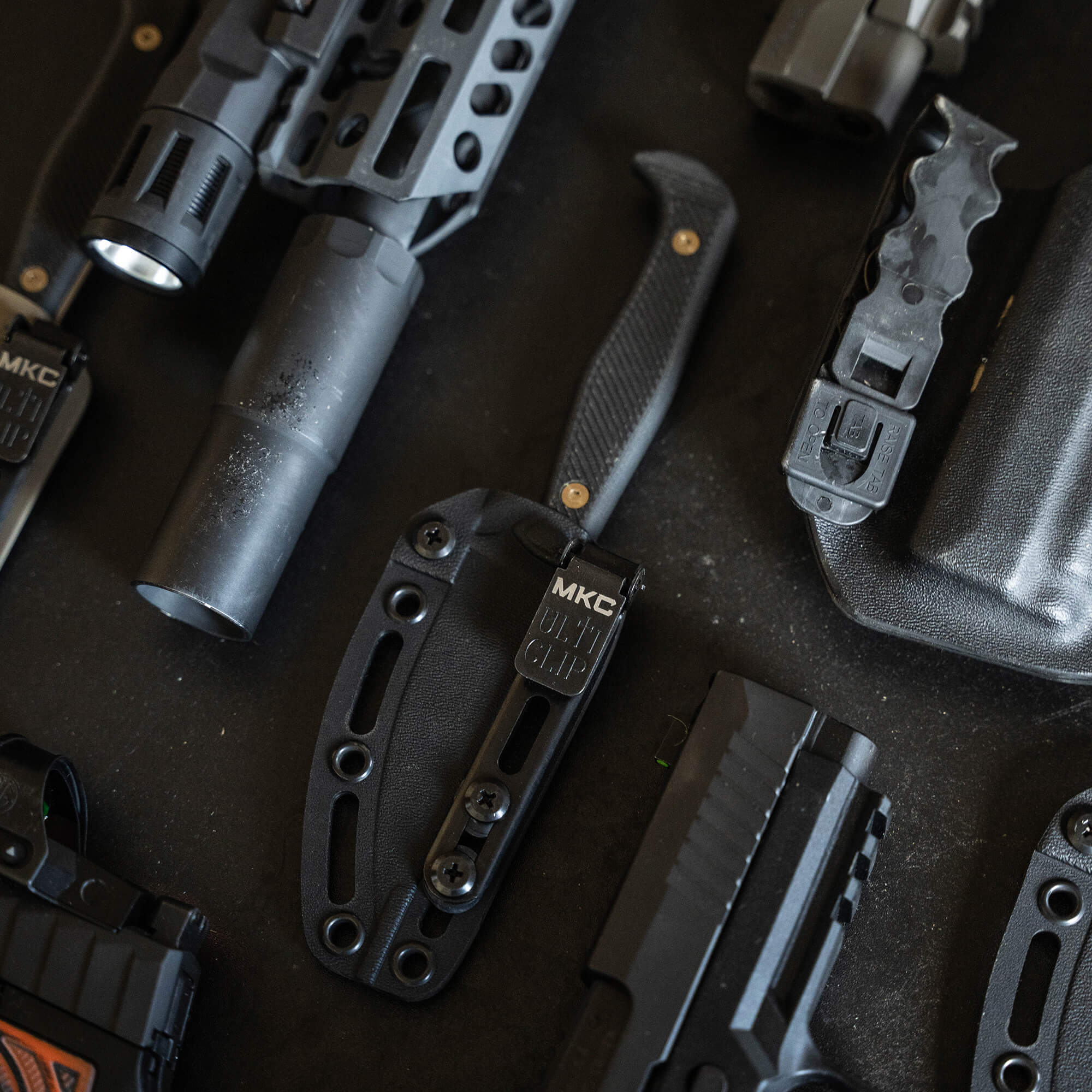Alt text: Assortment of tactical gear including knives, flashlight, and holsters displayed on a dark background, primarily in black, emphasizing a rugged and durable design.