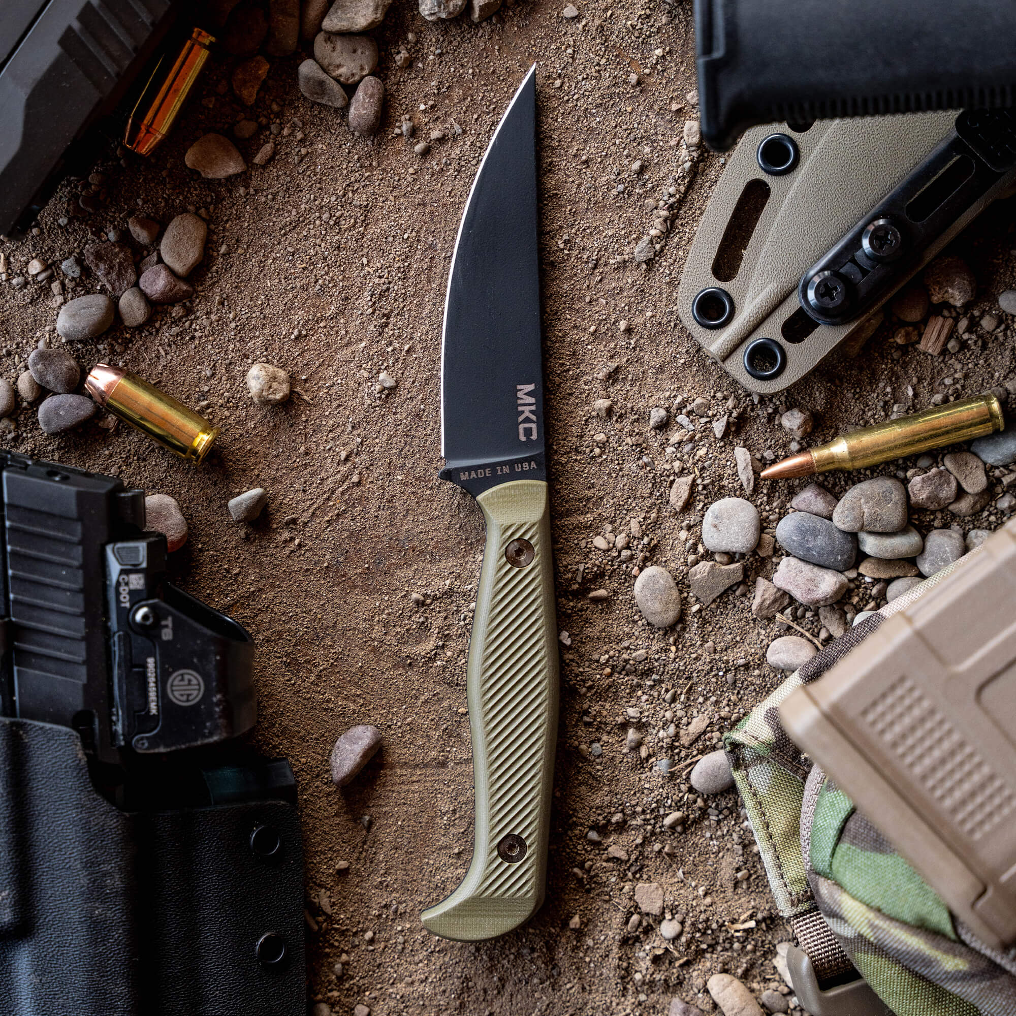 Alt text: "BATTLE GOAT fixed blade knife by MKC with black blade and olive drab handle, displayed on a sandy surface surrounded by tactical gear including a firearm magazine, spent bullet casings, and holster."