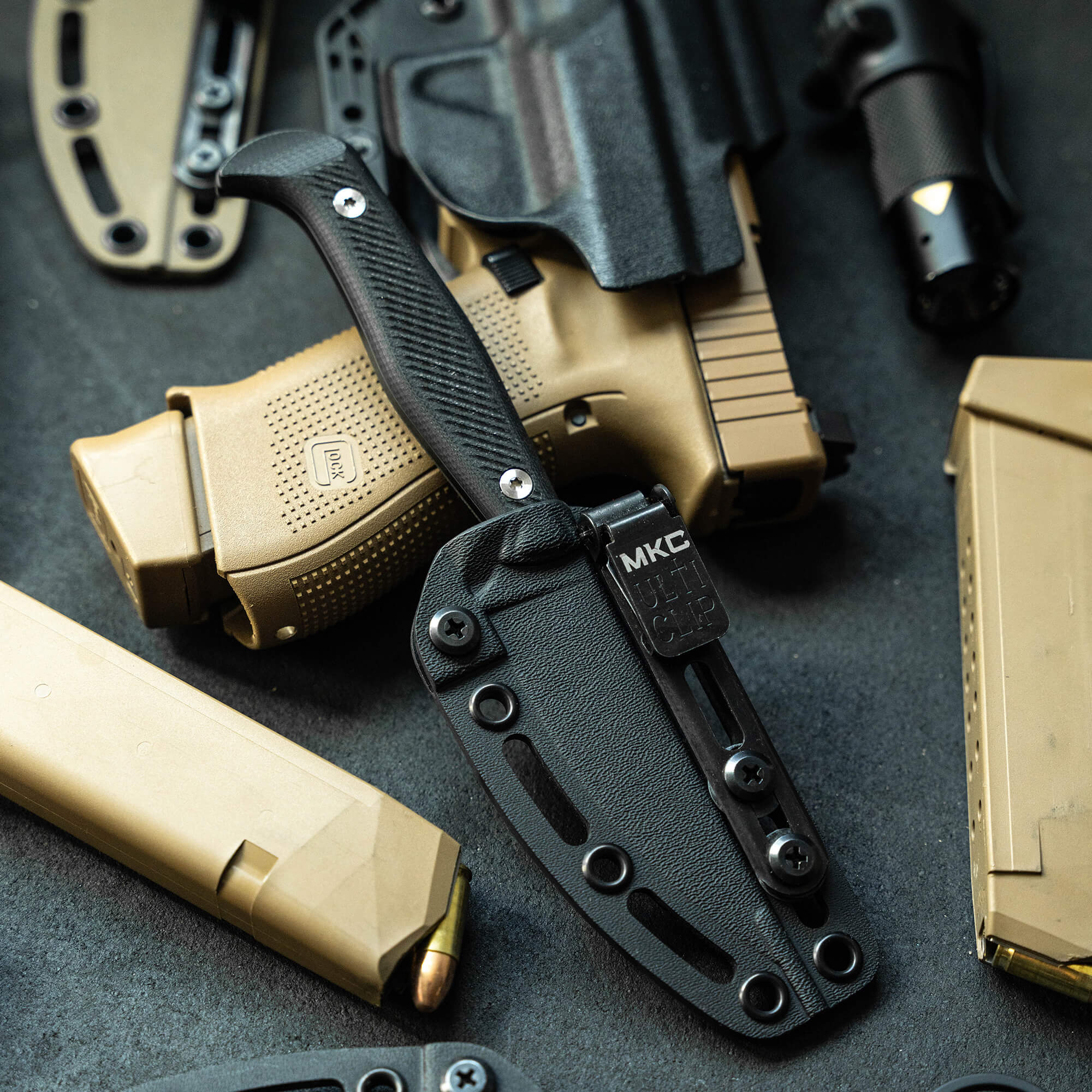Alt text: Close-up of a tactical knife secured in a black sheath, displayed alongside other tactical gear including a flashlight, holster, and ammunition magazines, all set against a dark gray background.