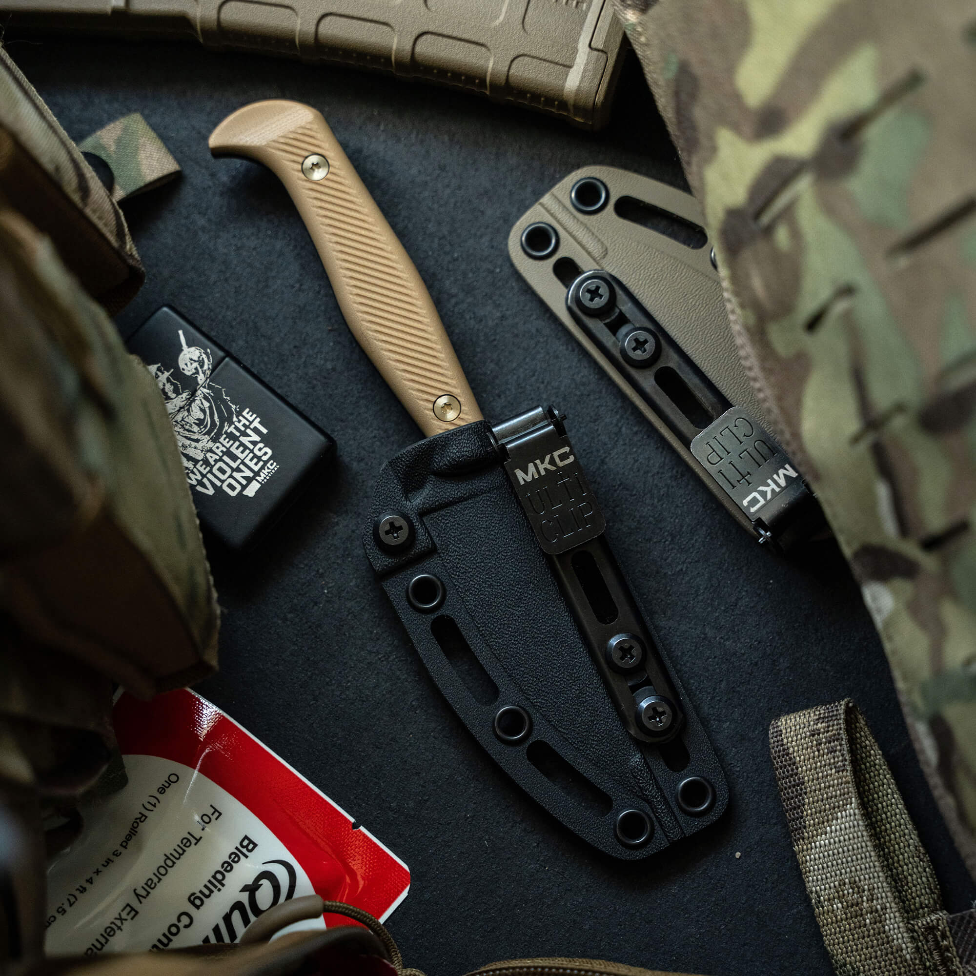 Alt text: "Close-up view of the Battle Goat tactical knife in black and coyote color, featuring a tan handle and black sheath, displayed among military gear including camo fabric, a tactical belt, and survival equipment."