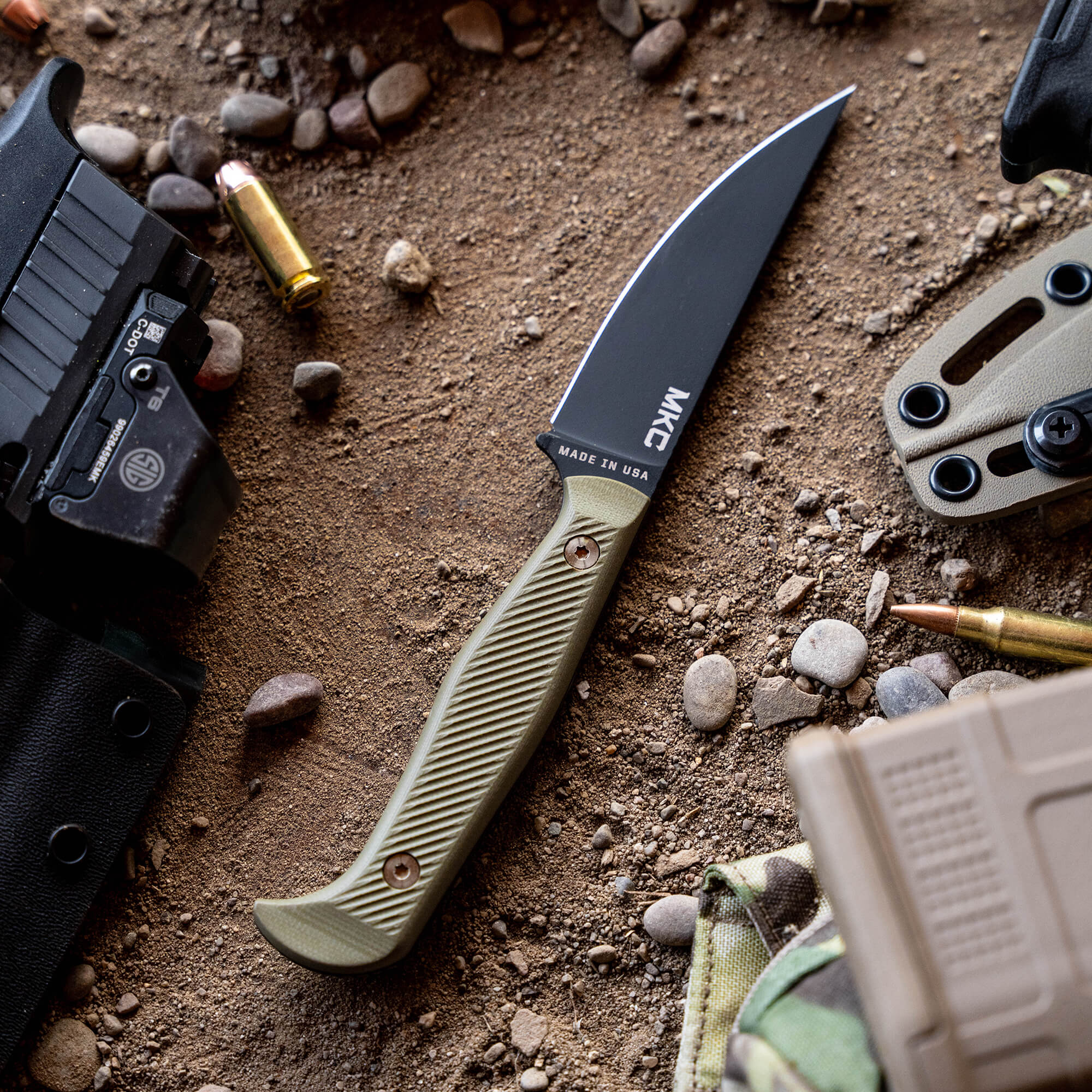 Alt text: "Close-up of the Battle Goat tactical knife with a black blade and olive drab handle, marked 'Made in USA', laid on ground amidst tactical gear, bullets, and rocky soil."