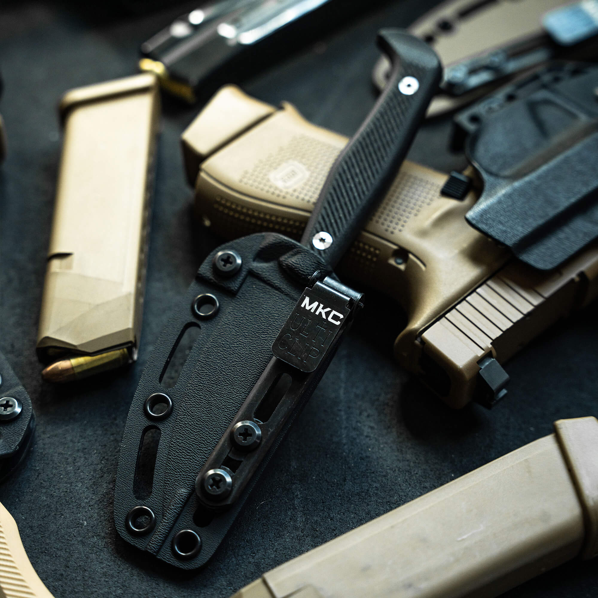 A close-up image of tactical gear including a black folding knife with a textured handle, labeled MKC, sheathed in a black holster, alongside other military-style items including a tan handgun and magazines.