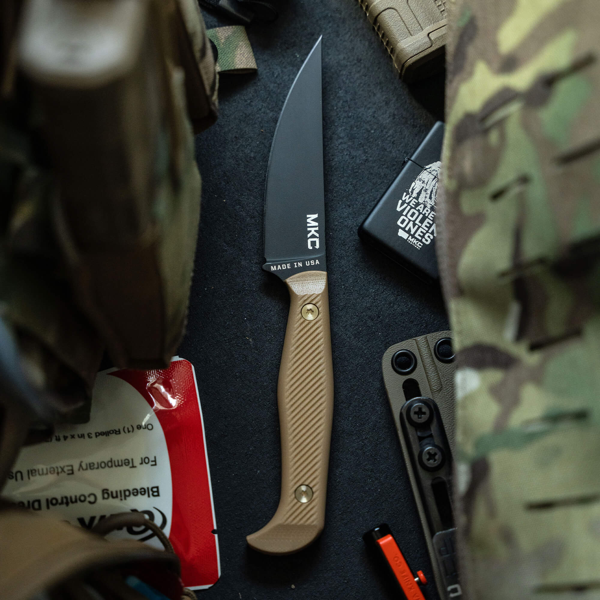 Close-up view of the 'Battle Goat - BLK/COY' tactical knife with a tan handle and black blade, surrounded by outdoor gear, showcasing its military-grade design and 'Made in USA' label.
