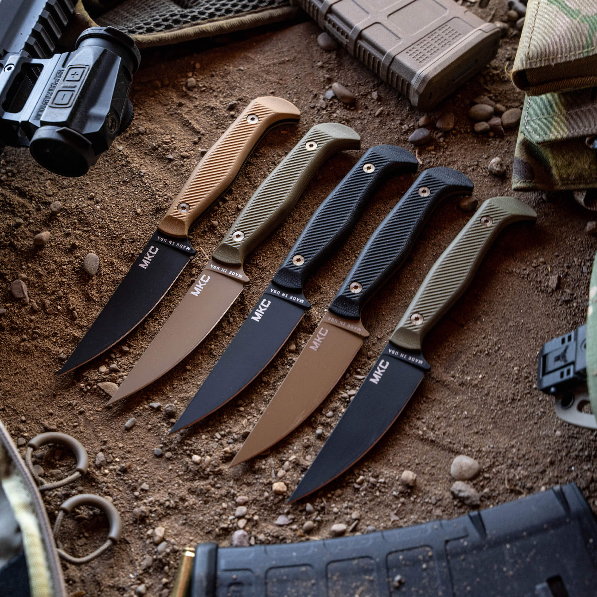 Alt text: "Array of tactical knives branded 'MKC' laid out on gritty surface with olive green, tan, and black handles, featured alongside tactical gear like a flashlight and a muddy tactical vest."