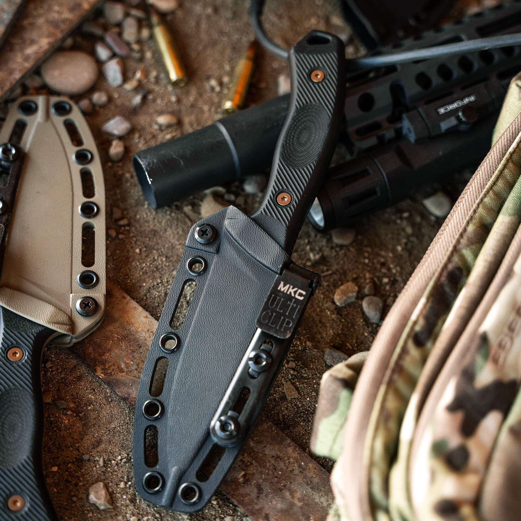 Alt text: "Close-up view of a black TF24 Kydex sheath with an MKC multi clip attached, displayed on a rustic background with a tactical rifle, ammunition casings, and an earth-toned bag."