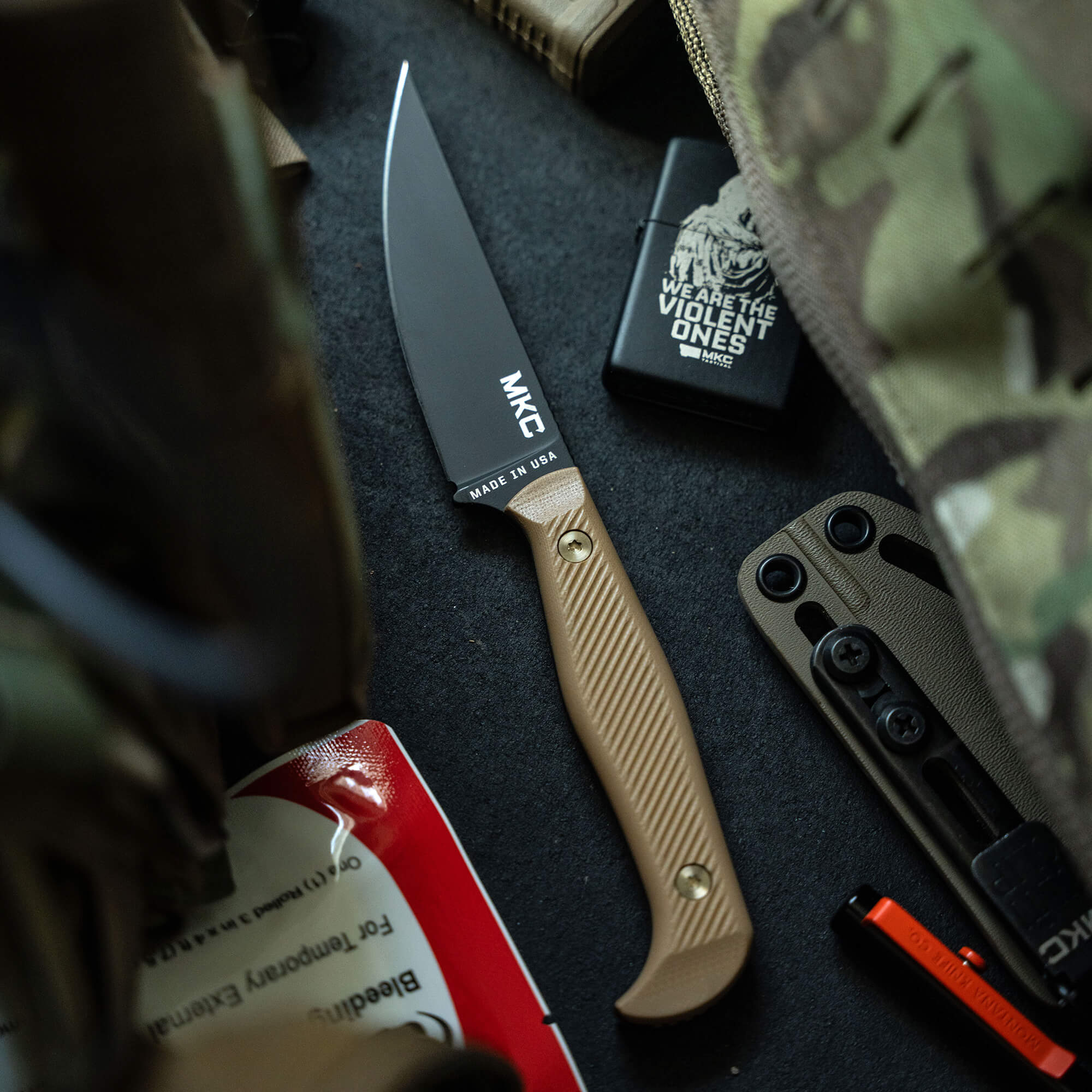 A tactical knife with a black blade and coyote brown handle, labeled "MKC" and "MADE IN USA," lying amidst survival gear including a camouflage backpack, tactical sheath, and a lighter.