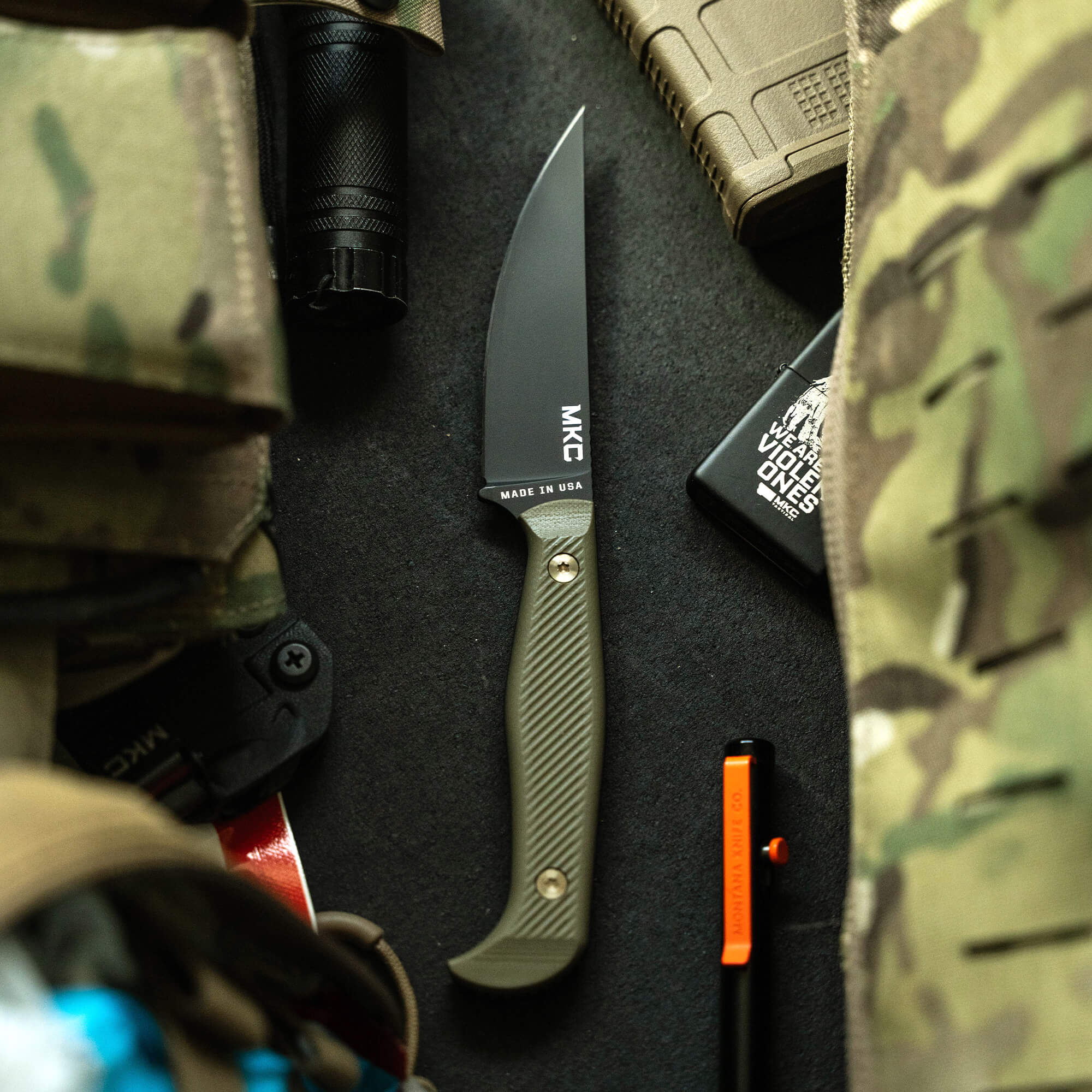 Black and olive drab MKC Battle Goat fixed blade knife with a textured handle, displayed on a dark, textured surface surrounded by tactical gear including a flashlight and camouflage material.
