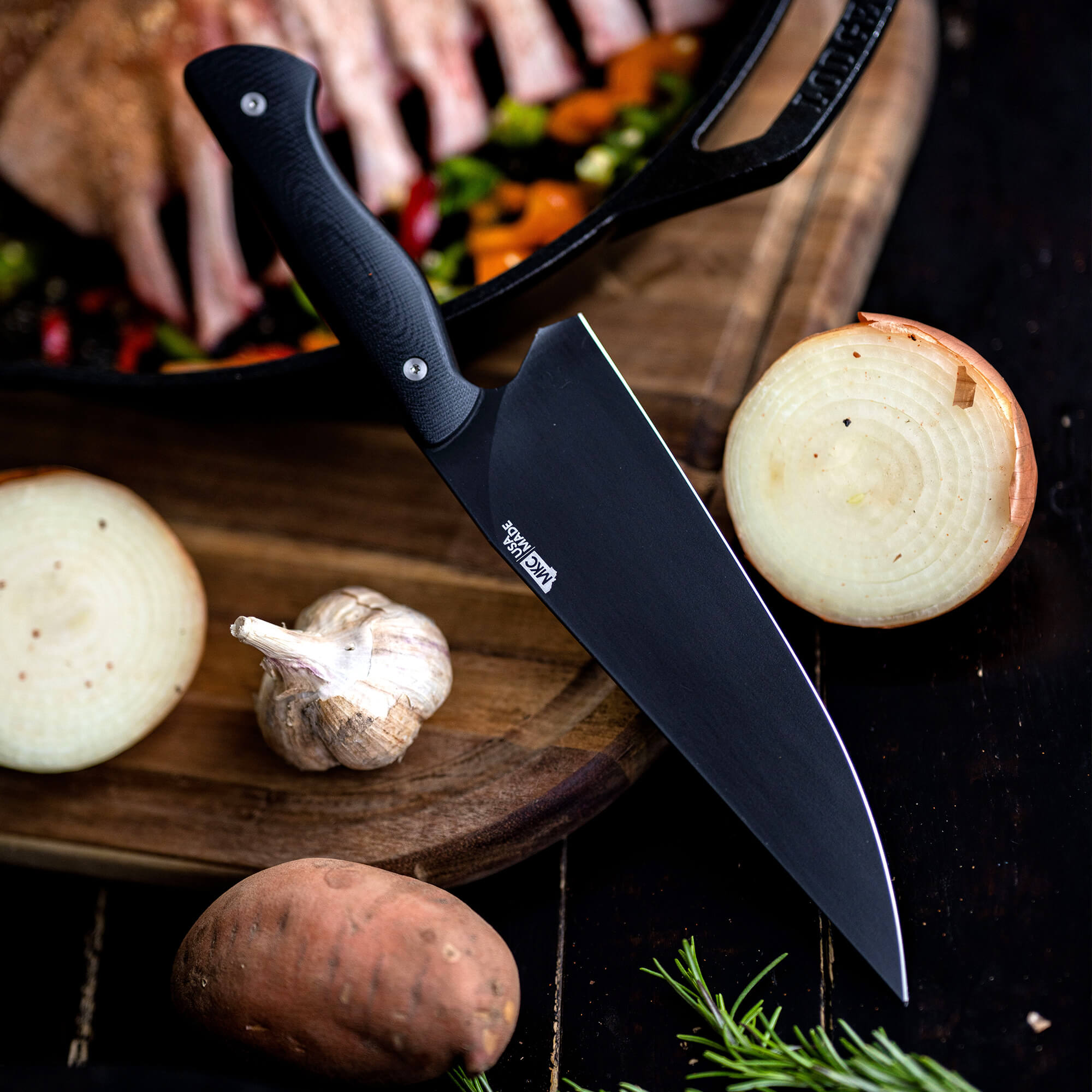 Alt text: "Close-up view of a Bighorn Chef black knife with a textured handle, resting on a wooden cutting board surrounded by ingredients including onions, garlic, and sweet potatoes, with a cast iron skillet loaded with herbs and meats in the