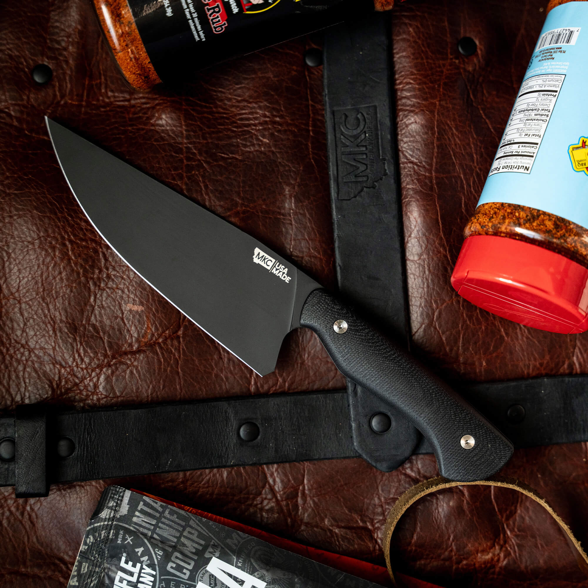 Alt text: "Meat Church chef knife with a black handle and silver blade, displayed on a leather surface alongside a black knife sheath, a Meat Church BBQ rub container, and a cookbook."