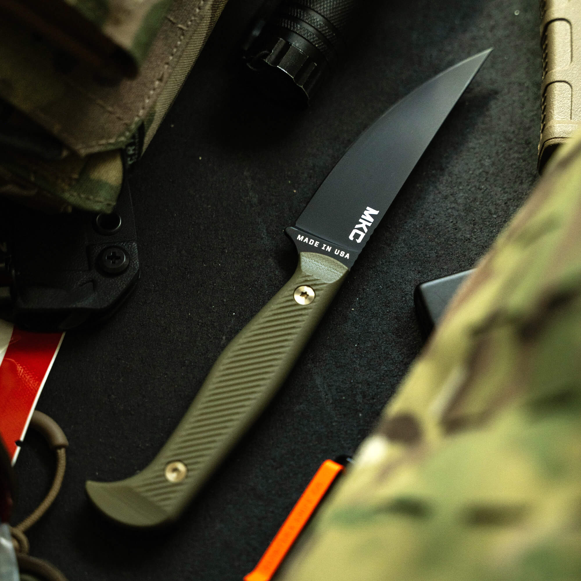 Alt text: "Close-up view of a sleek BATTLE GOAT tactical knife with a black blade and olive drab handle, marked 'MADE IN USA,' lying next to military gear on a textured surface."