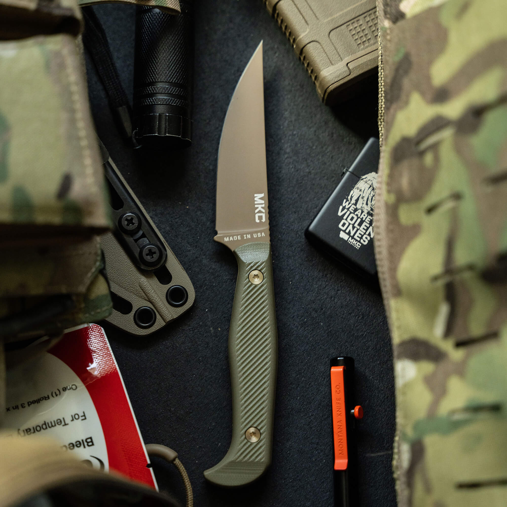 Alt text: "Tactical fixed blade knife with an olive drab handle and a tan sheath, prominently branded 'MKC' and noted as 'Made in USA', displayed among outdoor gear like a flashlight, boots, and camouflage material."