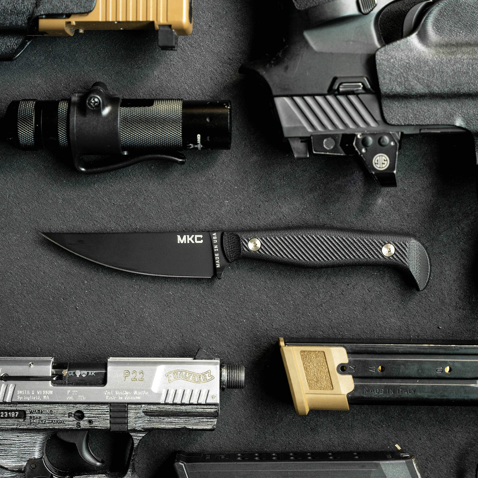 Alt text: "Close-up of a black tactical folding knife labeled 'MKE' on a textured handle, surrounded by various military-style firearms and equipment on a dark, textured surface."
