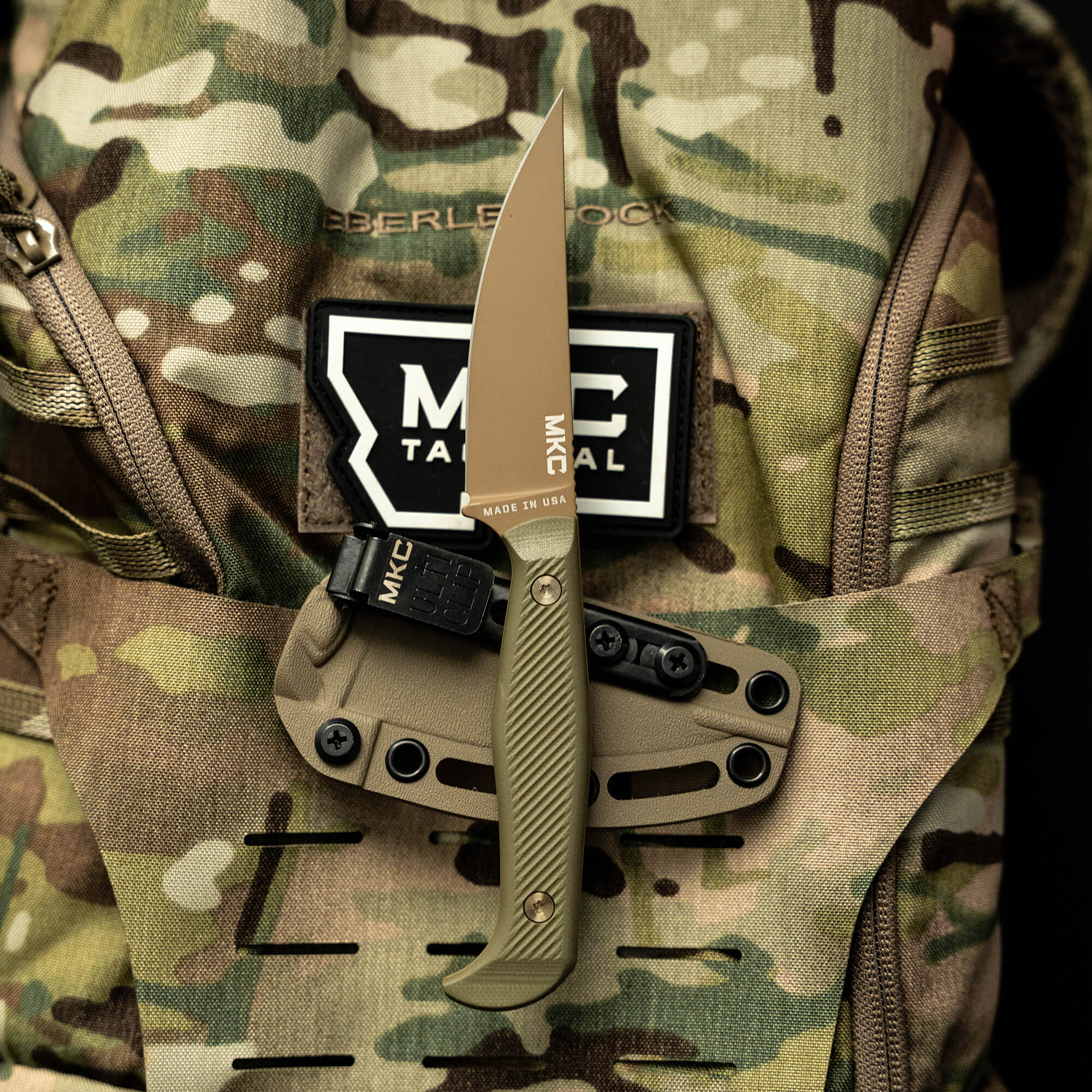 Alt text: "Close-up view of the Battle Goat tactical knife in coyote/olive drab color, displayed against a camouflaged tactical backpack with visible brand patch 'Berleben Rock.' The knife features a sleek, straight blade and