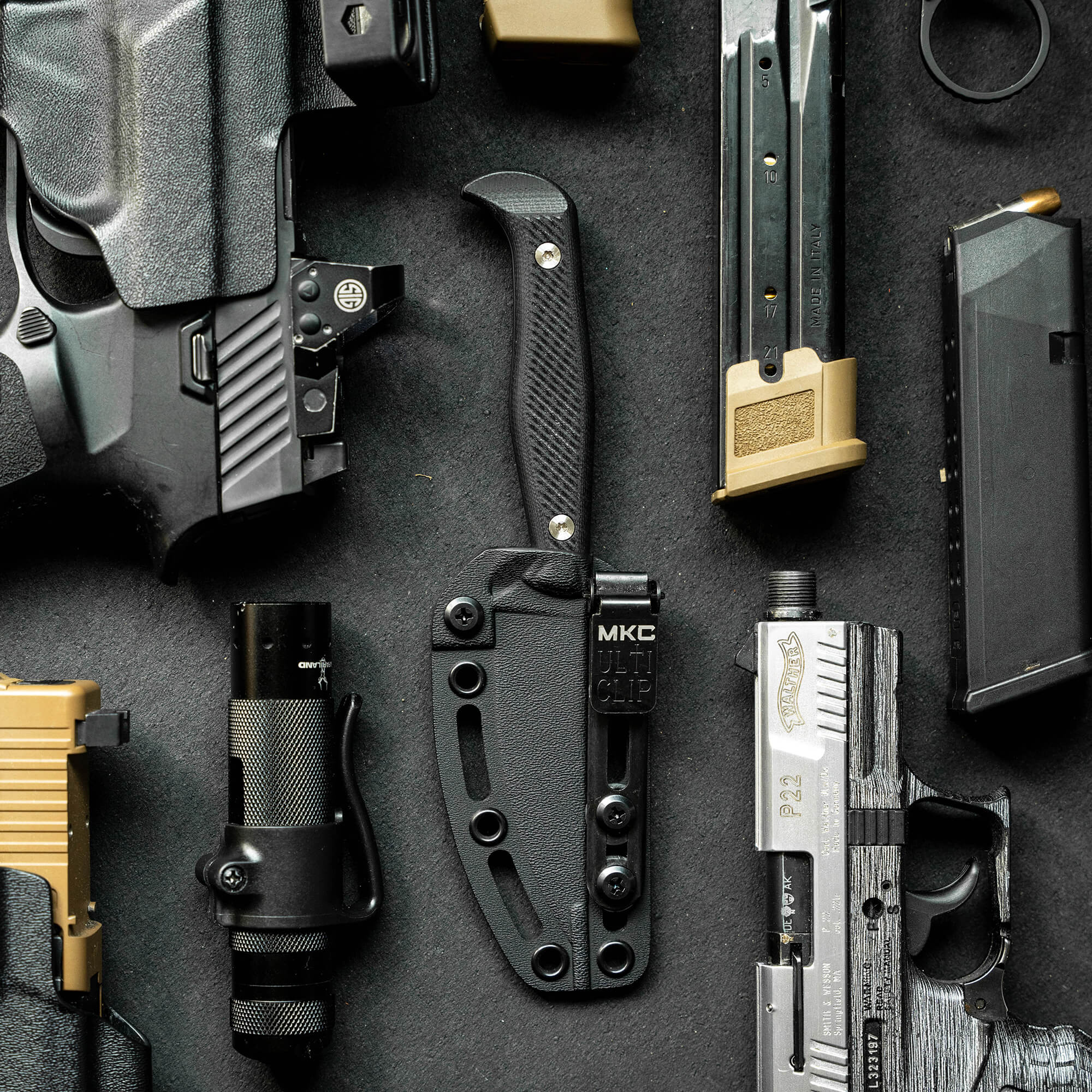 Alt text: "Product display featuring BATTLE GOAT tactical gear including a black knife, handgun, flashlight, holster, and various magazines, all arranged on a dark textured background."