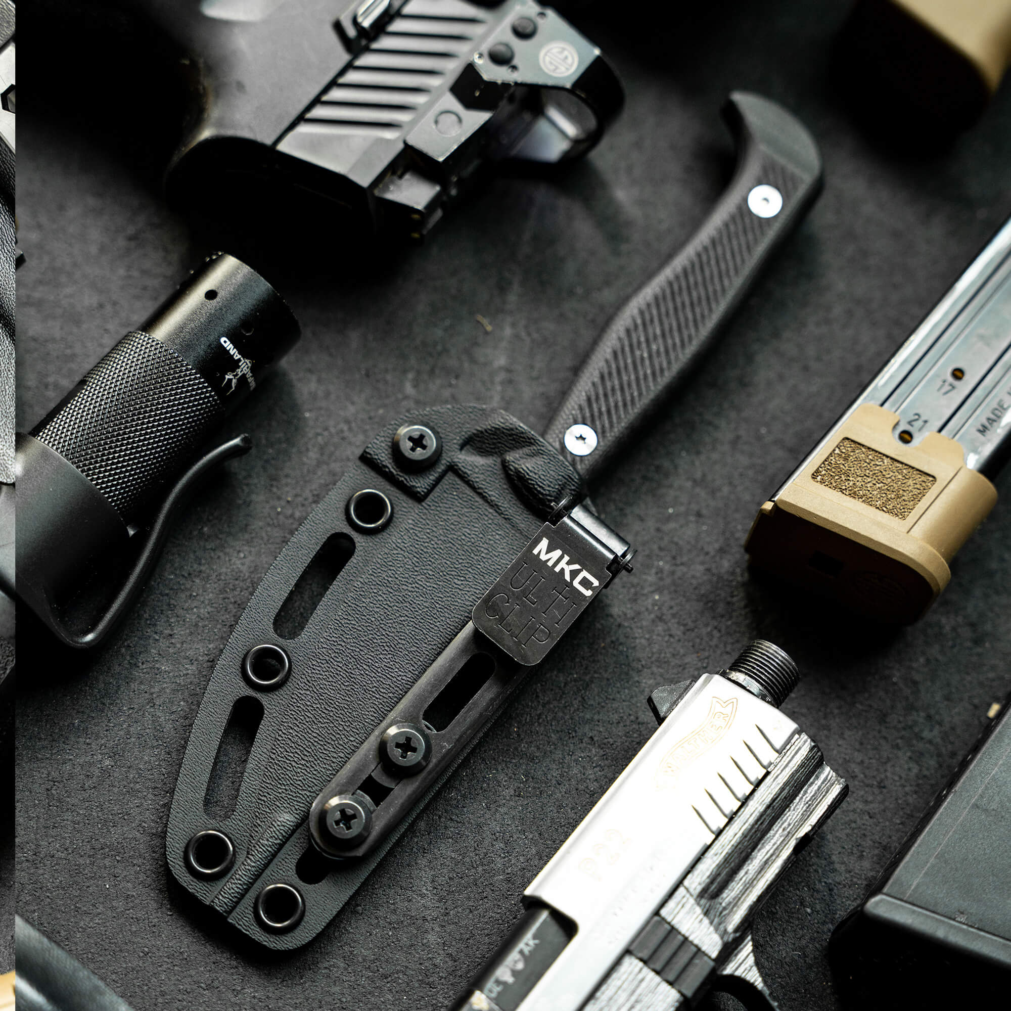 Alt text: "Close-up of a black tactical knife, BATTLE GOAT - BLK/BLK, sheathed in a black Kydex holster, surrounded by various tactical gear including magazines and a flashlight."