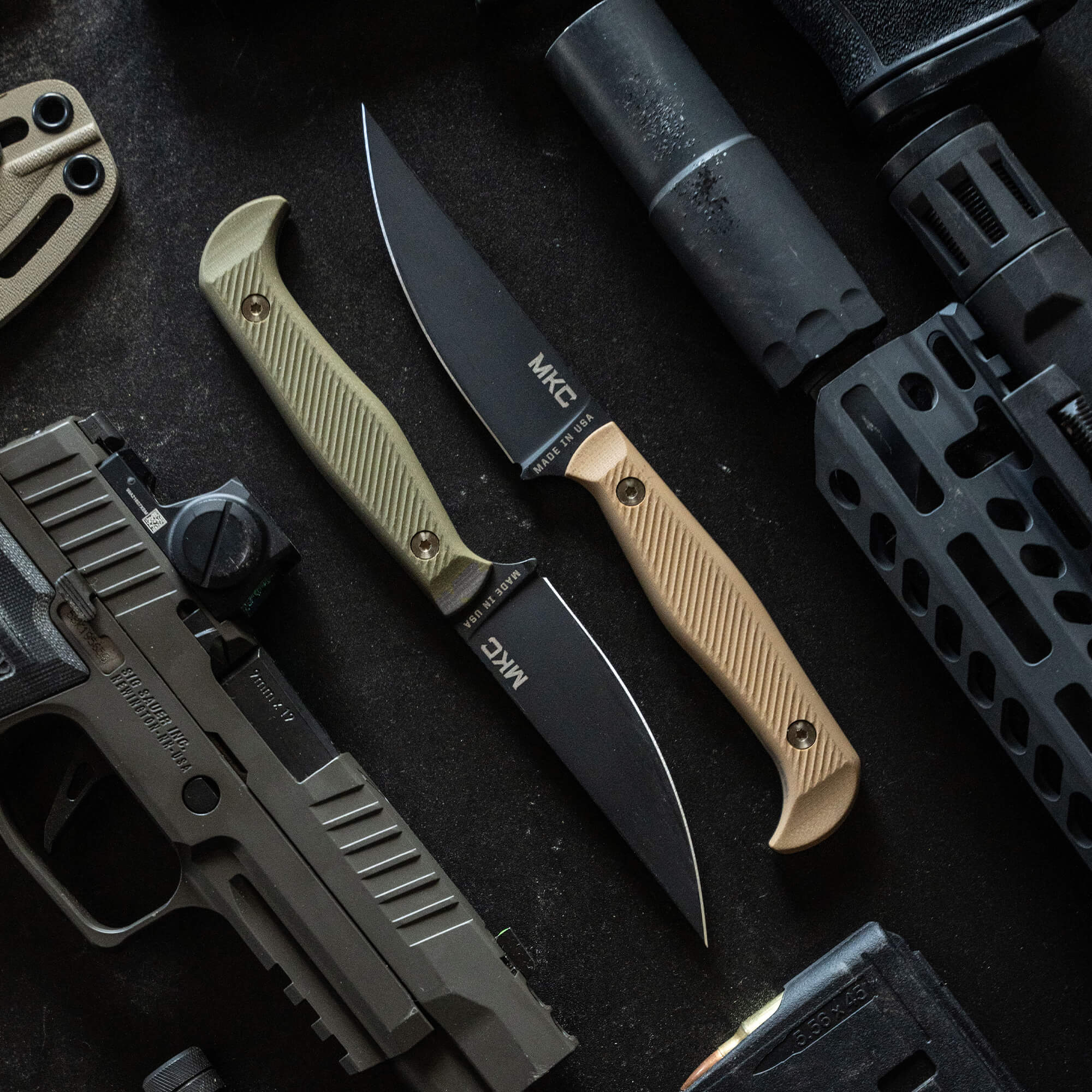 Alt text: "Top view of two BATTLE GOAT BLK/OD knives with tan handles, prominently displayed next to tactical equipment including a handgun, rifle parts, and a suppressor on a dark background."